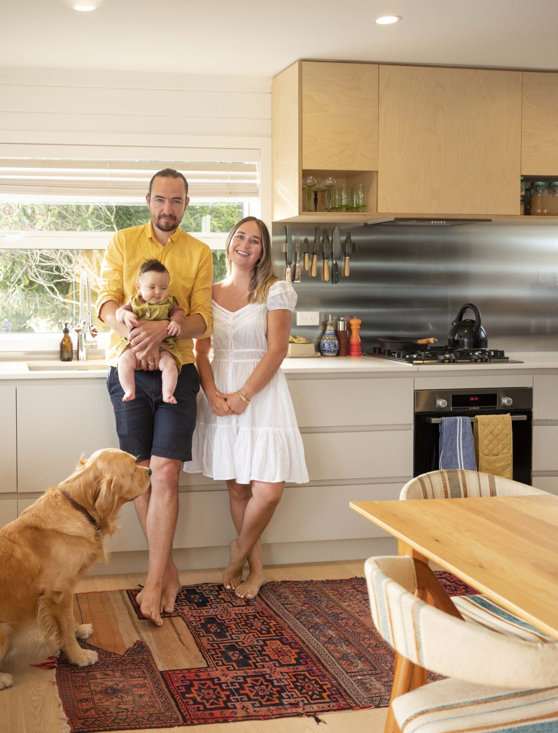 A couple stand in their kitchen holding a baby with their dog by their side