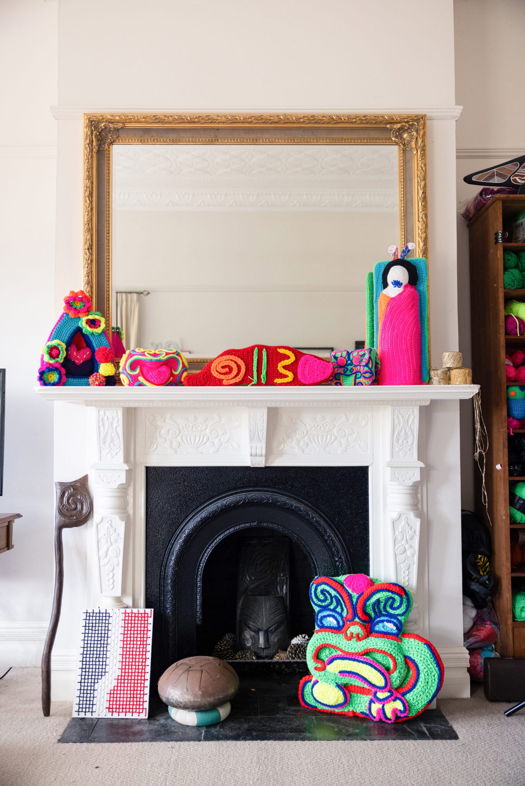 A white ornate fireplace with a large gold mirror on shelf and assorted crochet art around it