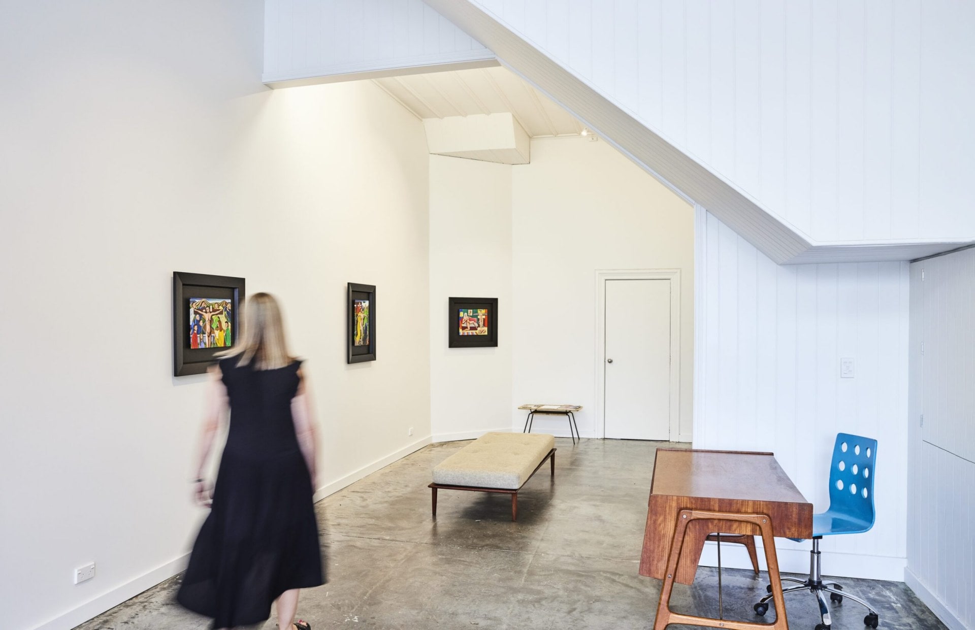 Interior of Suite gallery in Ponsonby. A woman wearing a black dress is walking through a tall white room with grey floors and three hanging artworks