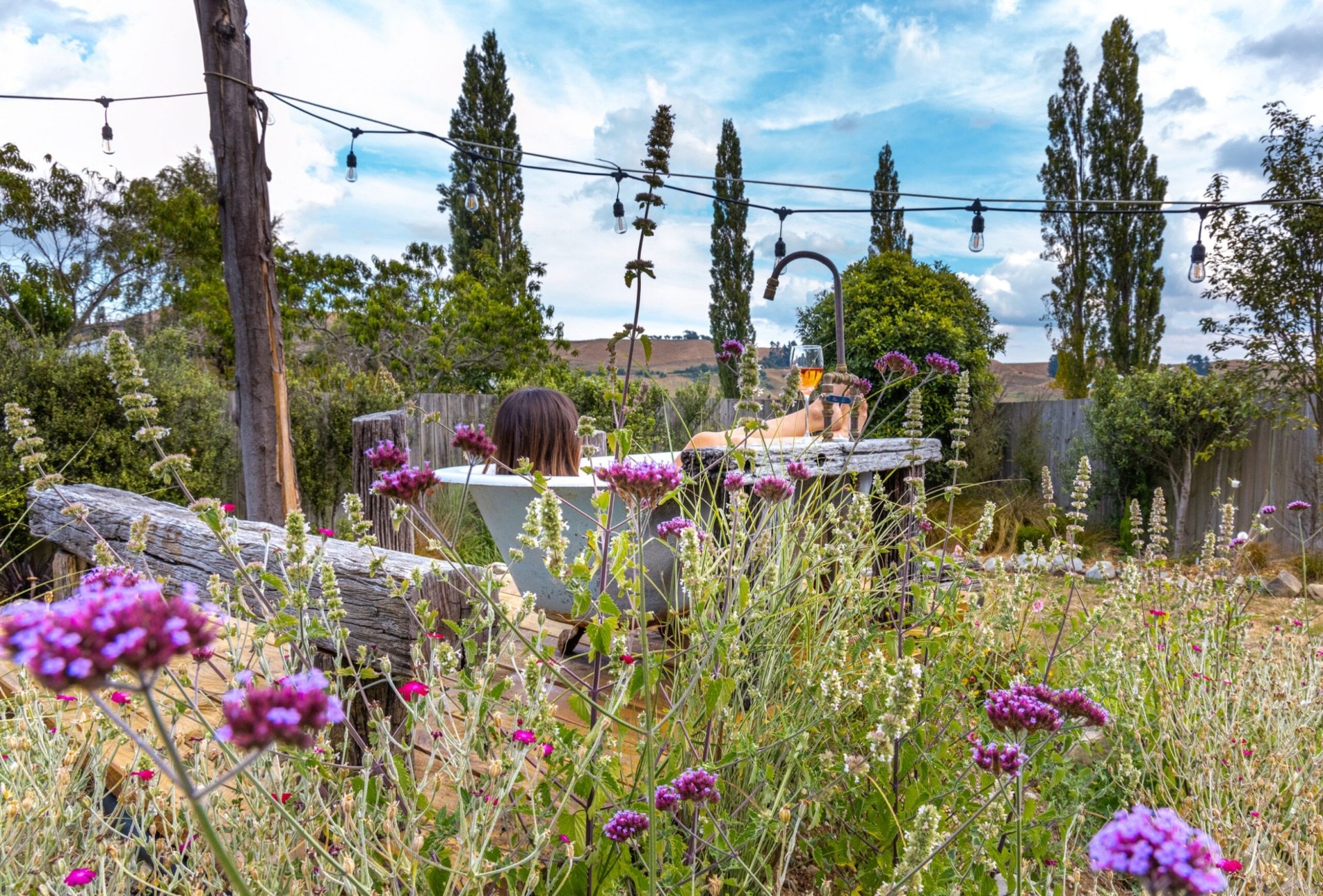 Woman sitting in a tub outside surrounded by tall grass and purple flowers