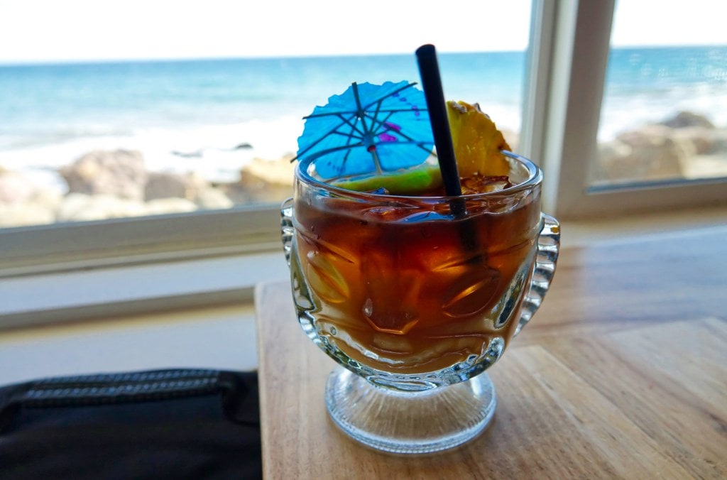 A Mai Tai cocktail with blue umbrella and pineapple slice in a classic tiki glass, with the Pacific Ocean in the background.