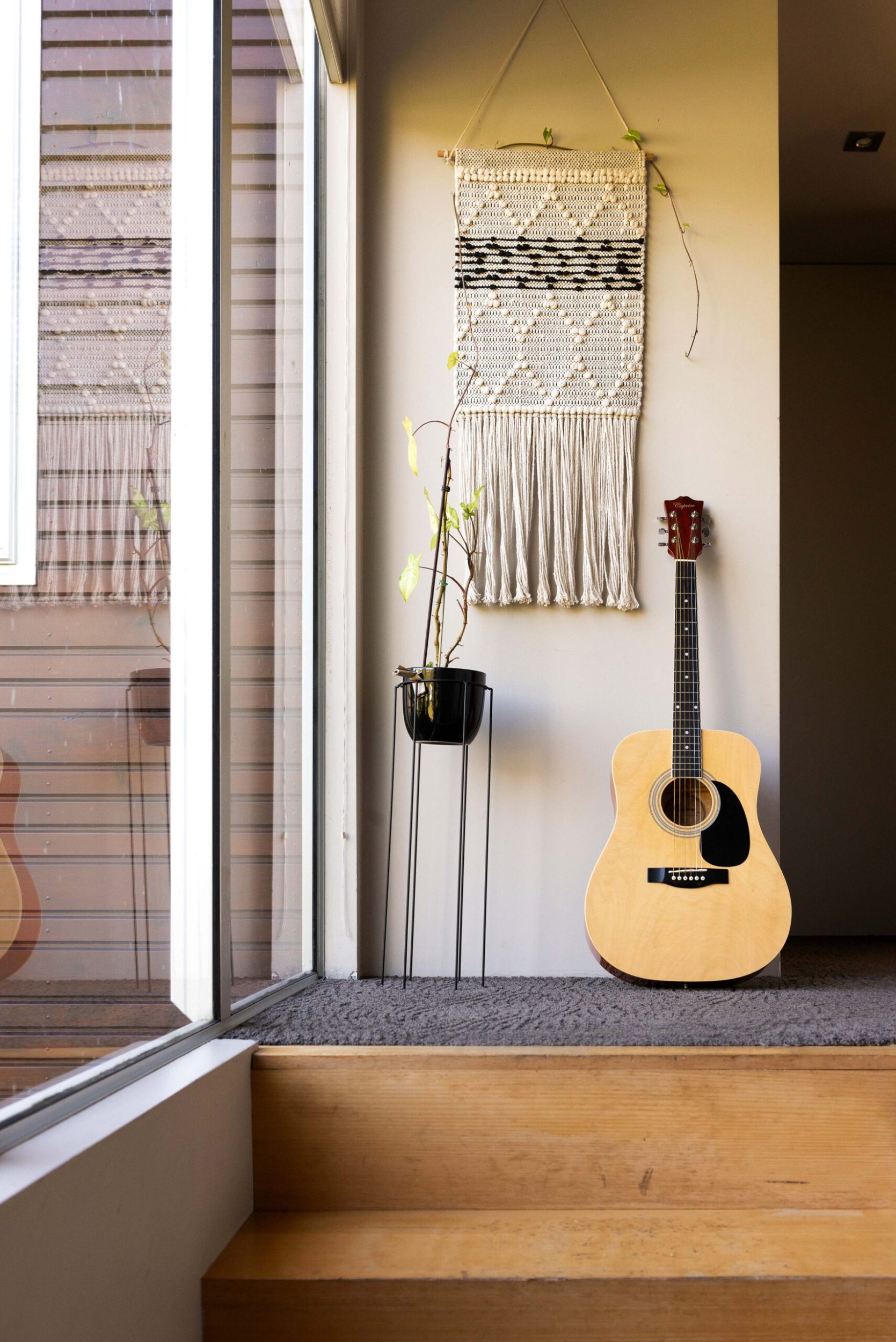 A guitar leaning against a white wall, with a hanging wall decor and pot plant next to it 