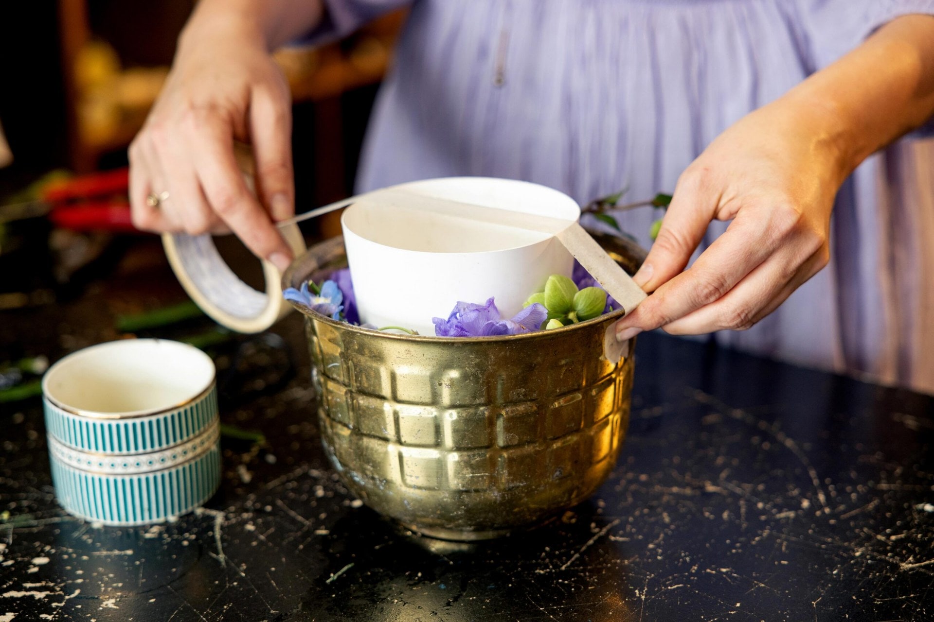 A woman in a purple dress taping a small bowl filled with rice to the larger metal bowl filled with flowers