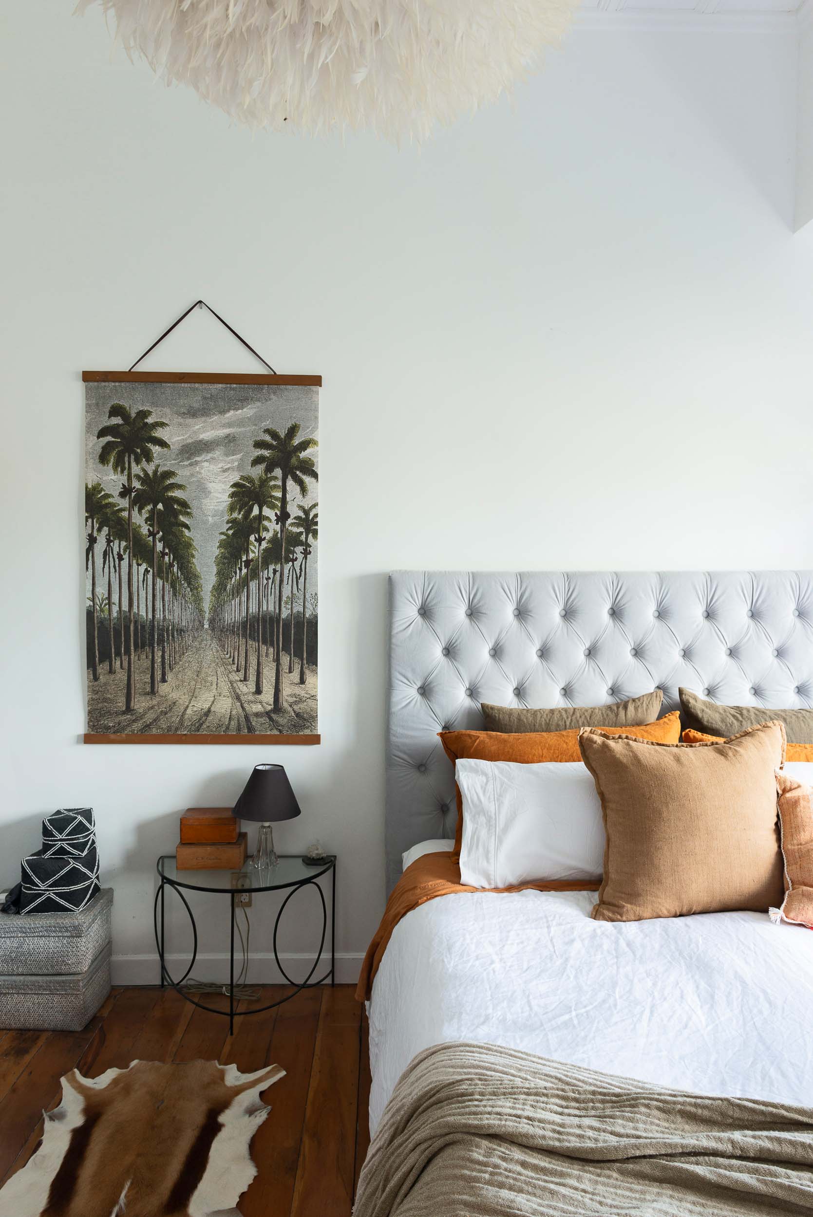 A bedroom with a bed with a light grey tufted headboard, a piece of palm art hangs on the wall and an iron and glass bedside table