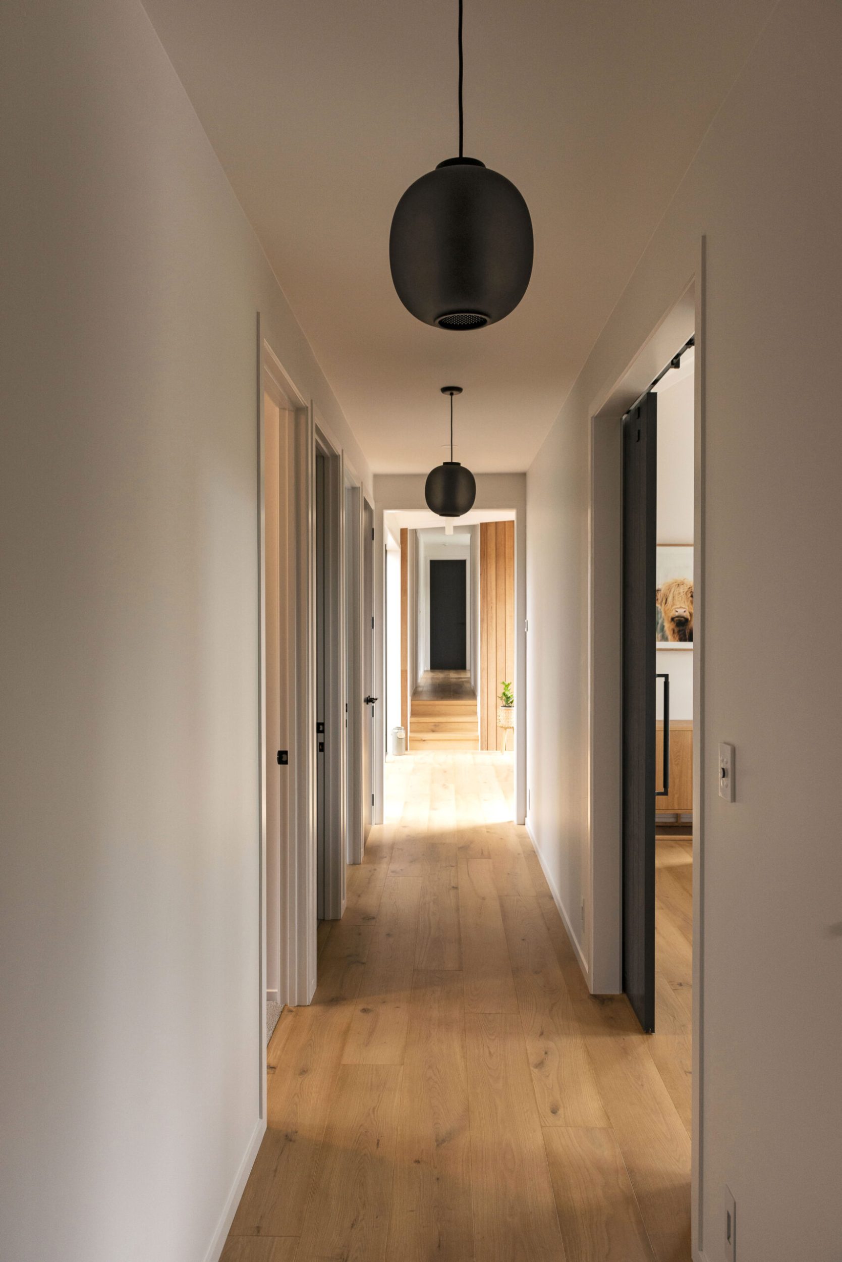 A long hallway with cream walls, wood floors, round black lamps 