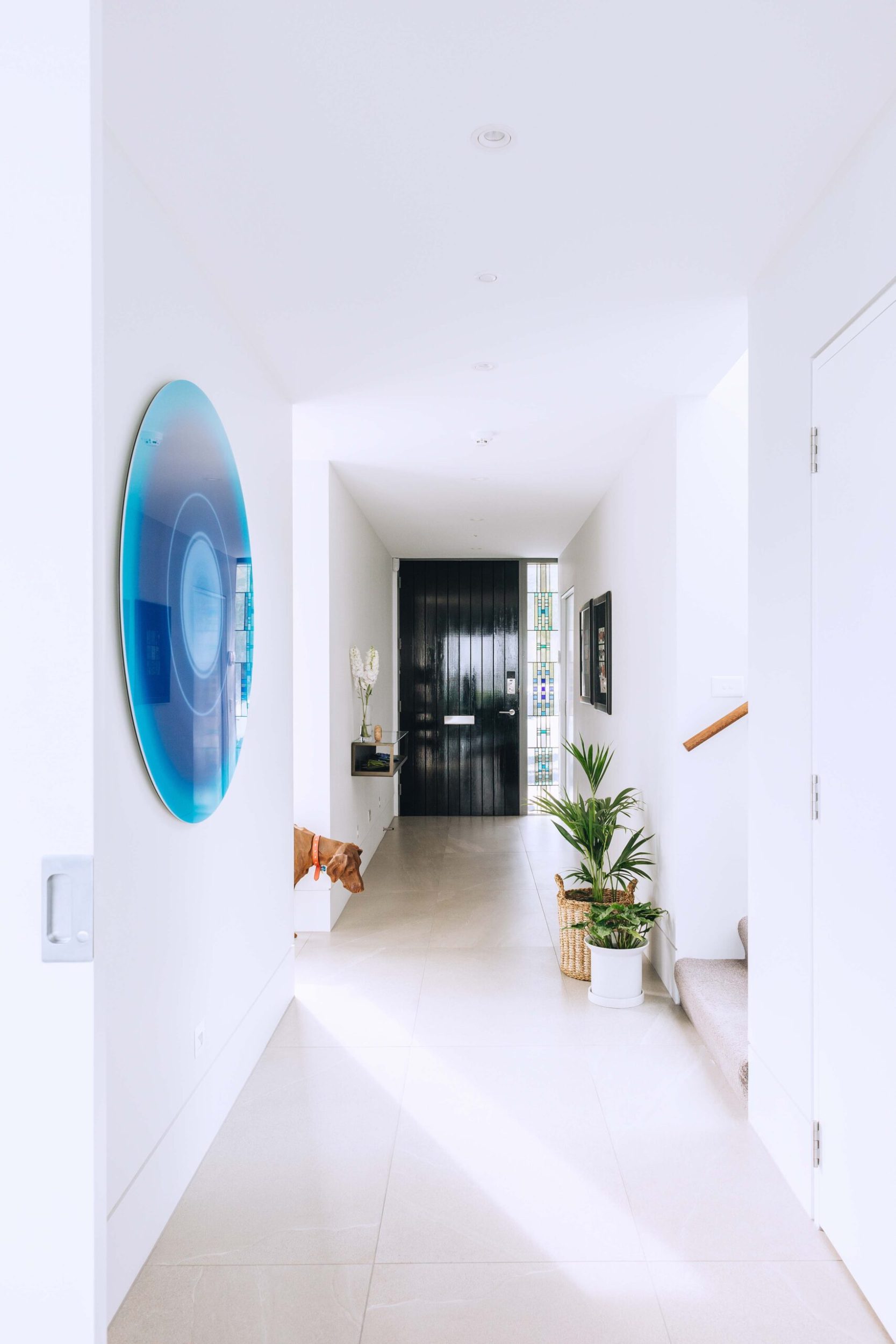 Home entryway with white walls, grey floor tiles, a black door and a round large blue decorative Max Patté wall art