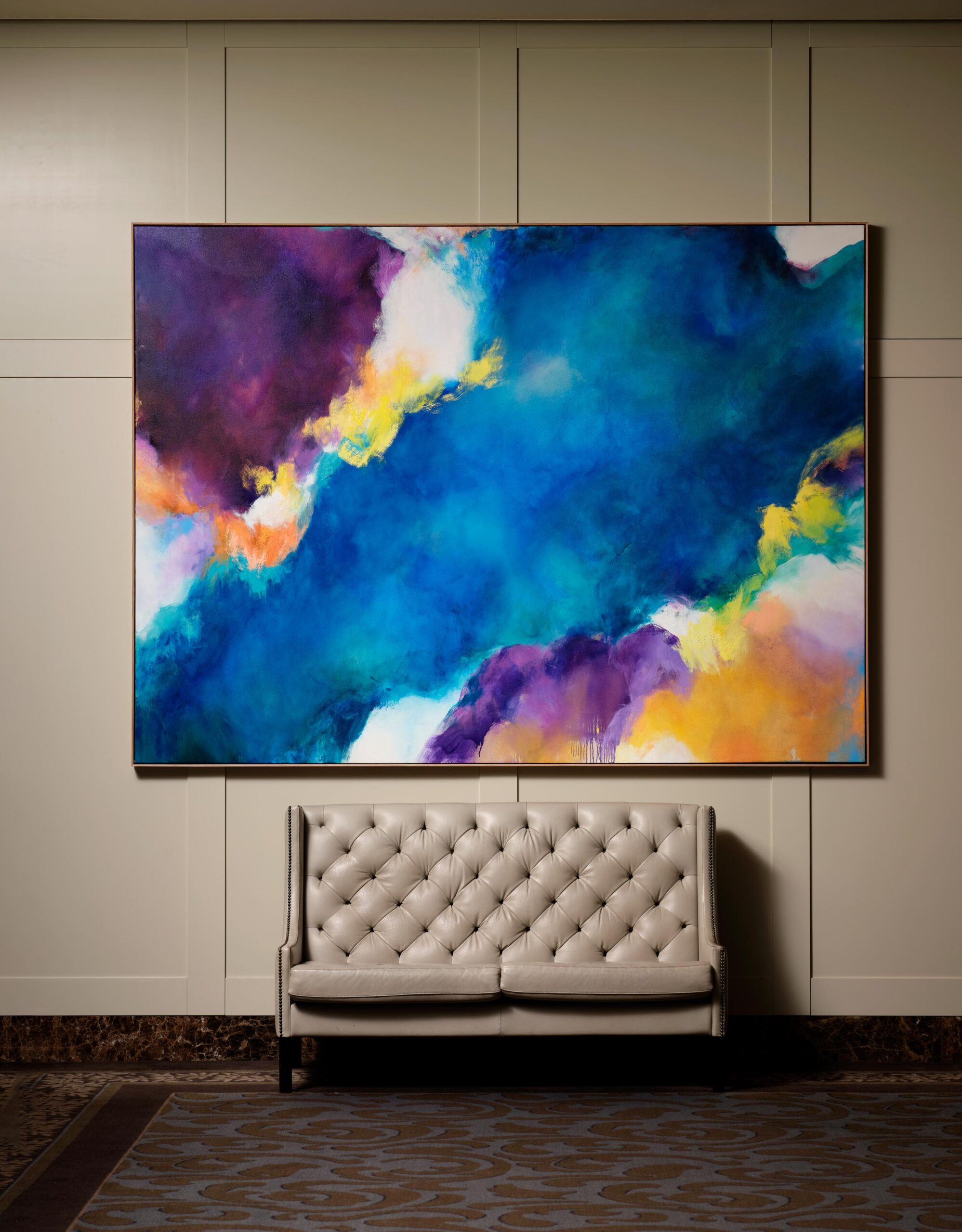 neutral toned small couch, that sits below a large abstract painting of blues, purples and yellow.