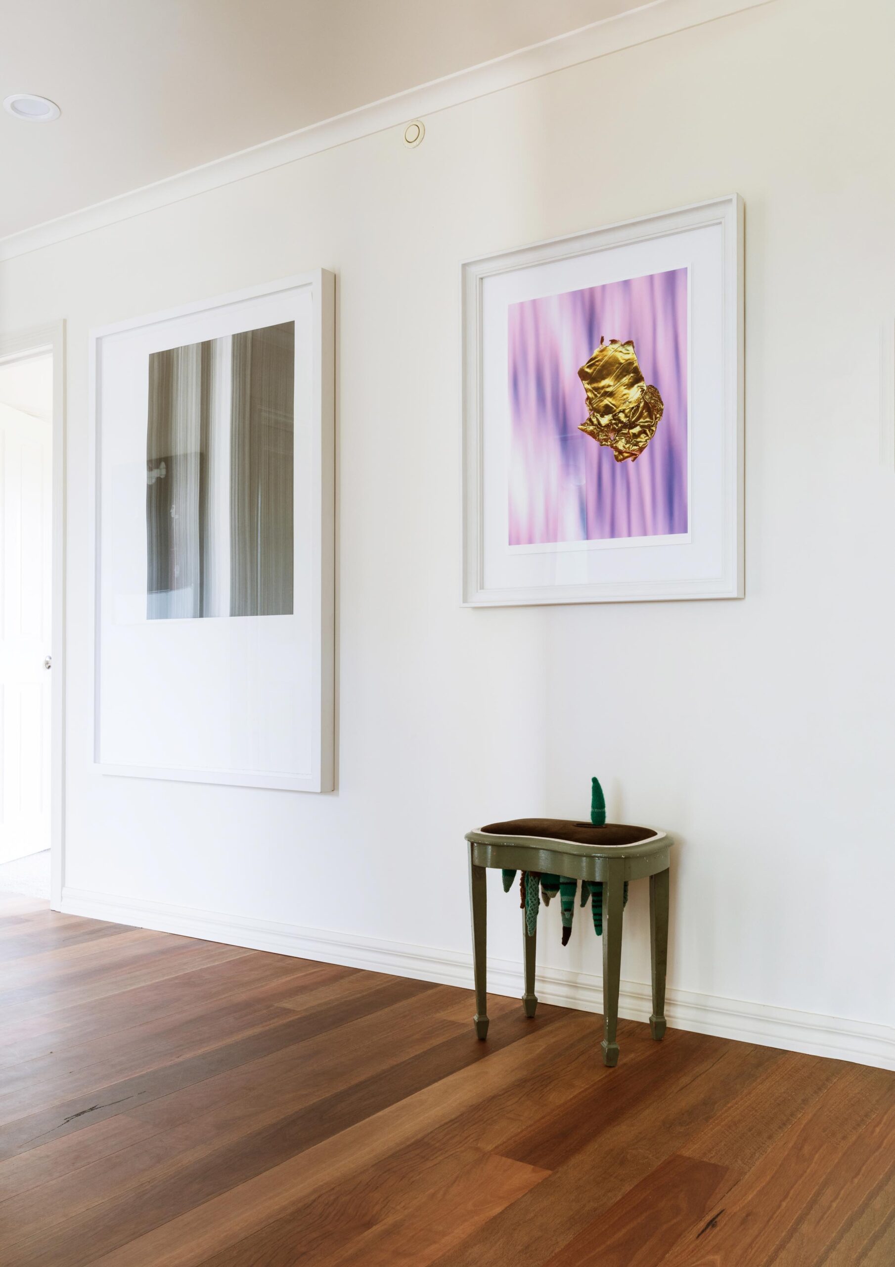 Two large pieces of art in white frames hang on a white wall with a vintage stool on the ground
