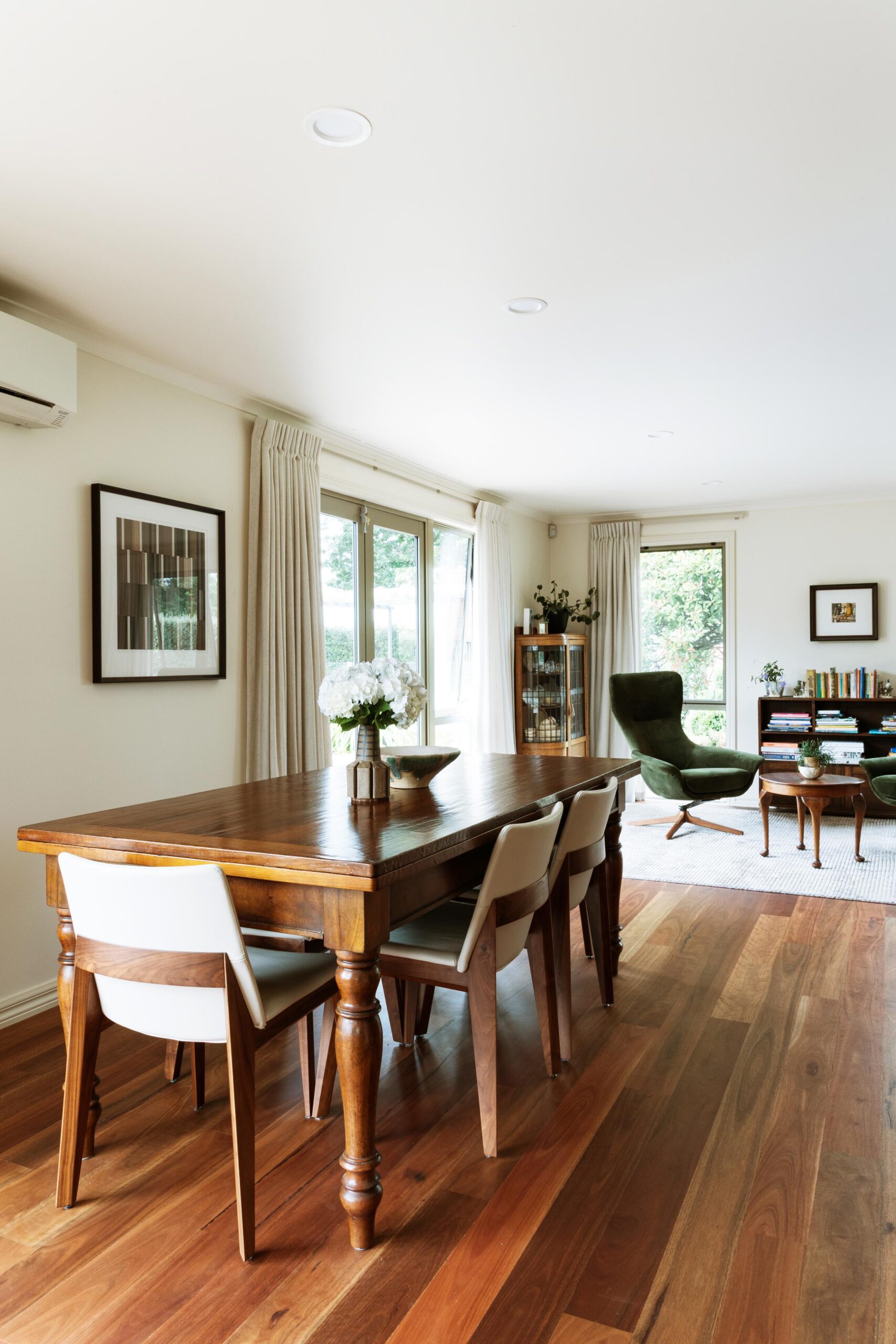 An antique dining table on hardwood timber floors, in the distance of the image is a green chair and an antique display cabinet