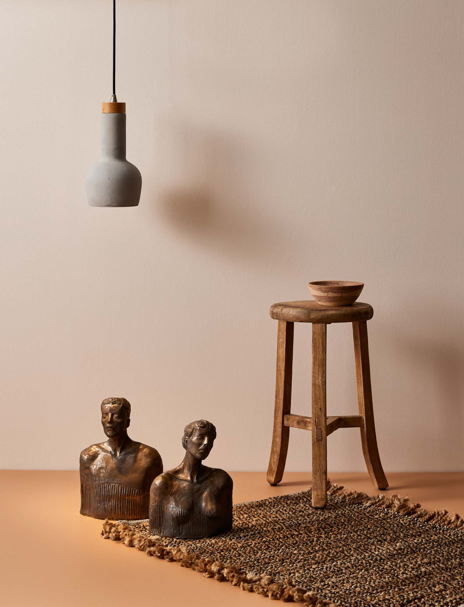 Image with beige wall and floor, a wooden stool with a bowl sitting on the stool, there is a jute rug on the floor and wooden bust statues on the rug