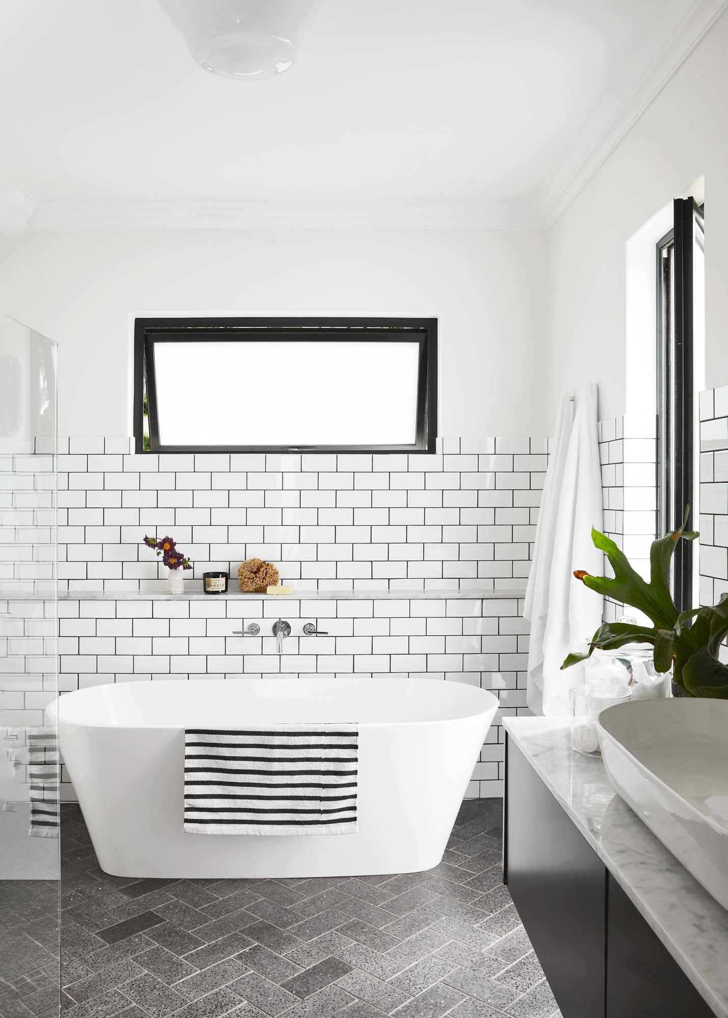 A bathroom with white subway tiles on the wall, a rectangular window above the tile line, a free standing bath in the middle, and grey subway tiles in a herringbone pattern on the floor