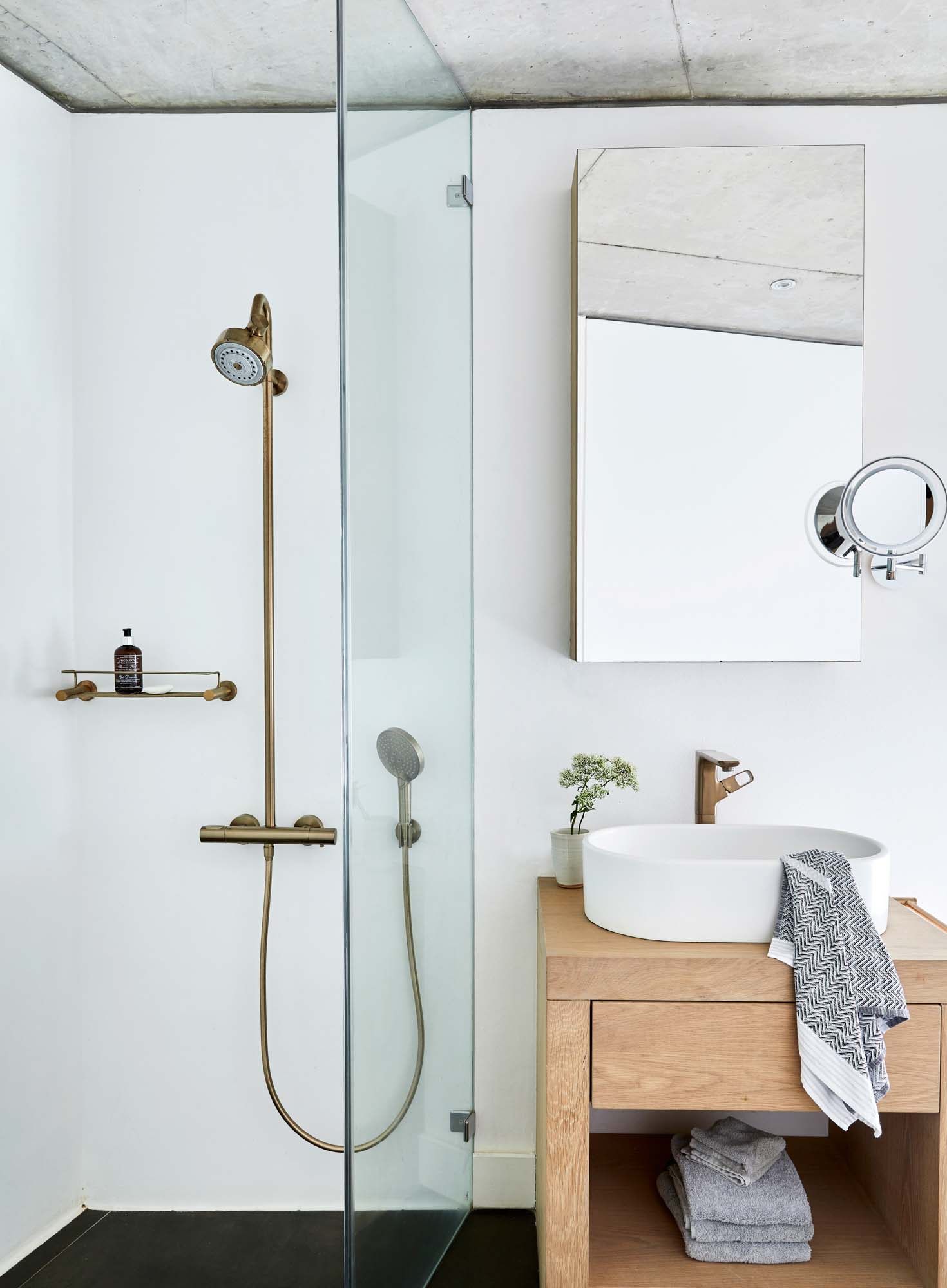 Bathroom with a shower to the left with a brass head and hose and glass shower screen. A timber vanity sits next to the shower with a single white sink