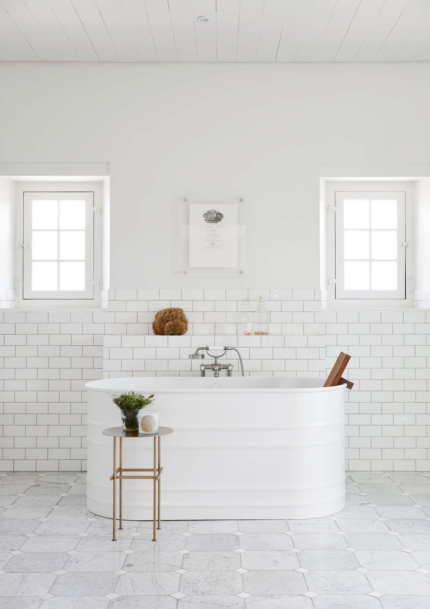 An all white bathroom with white subway tiles on the wall, marble white square tiles on the floor and a white metal bathtub in the middle of the room