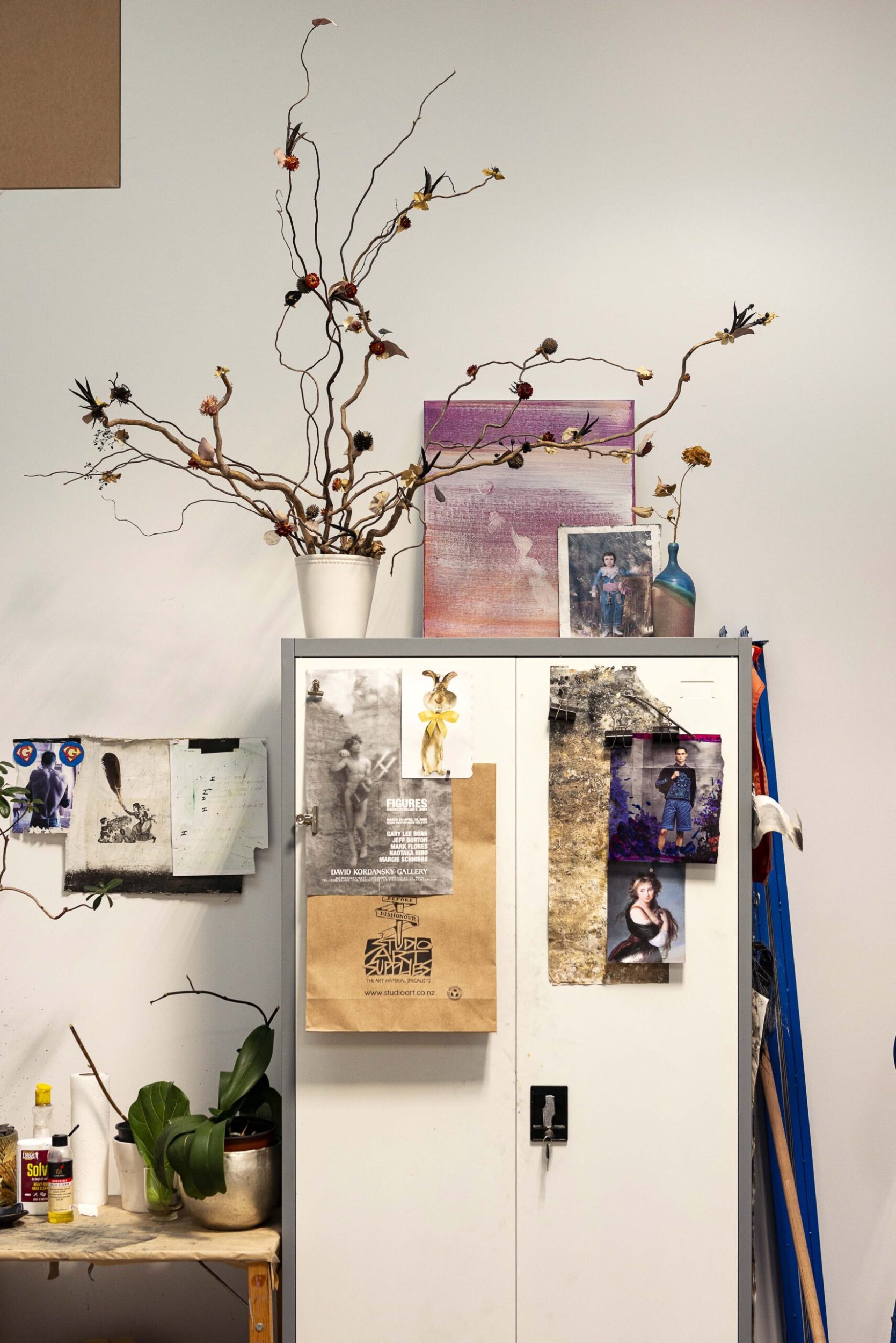 White storage cupboard with art pinned to it and a pot with dried floral arrangement placed on top