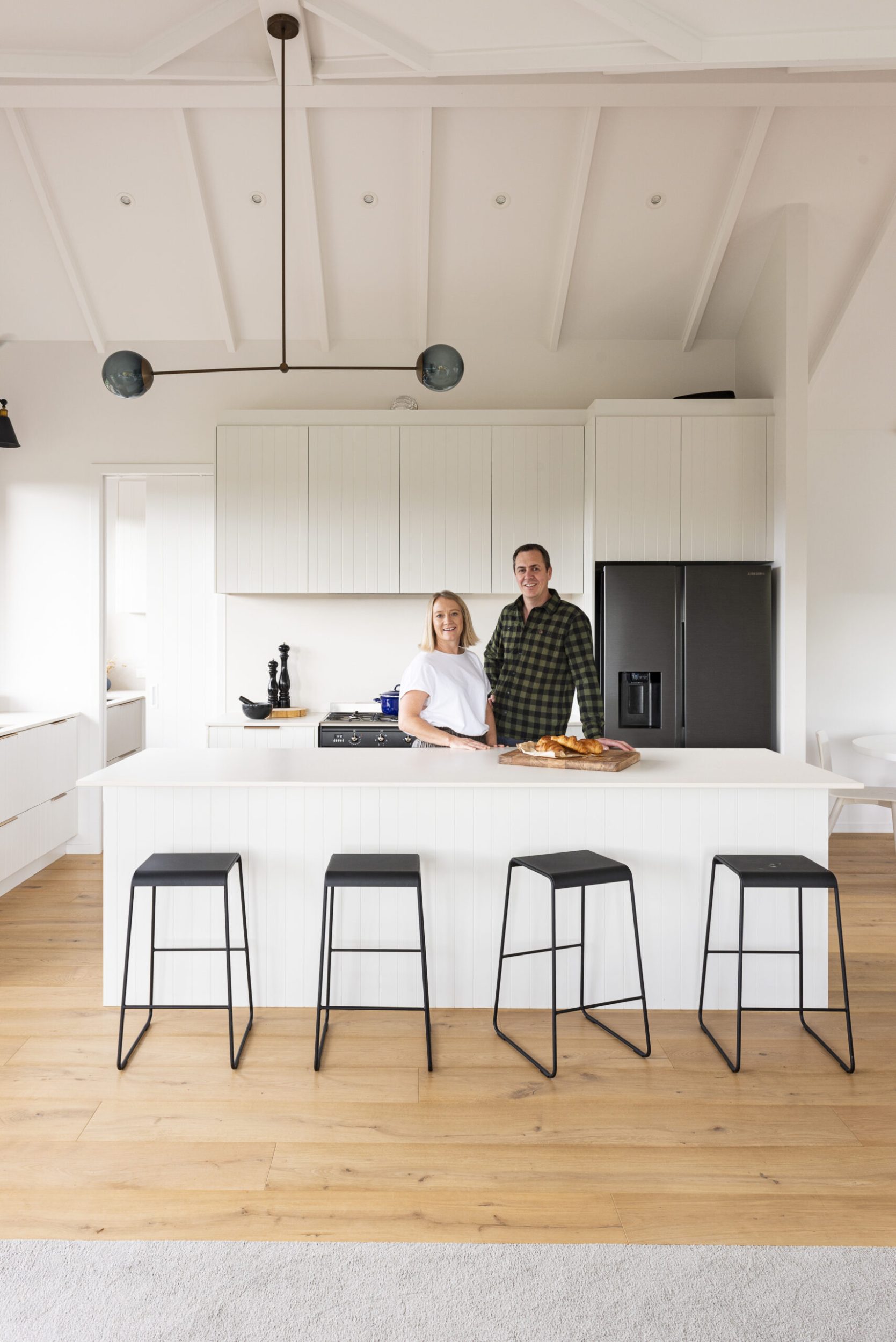 A white kitchen a white island, wood floors, a black fridge and black stools