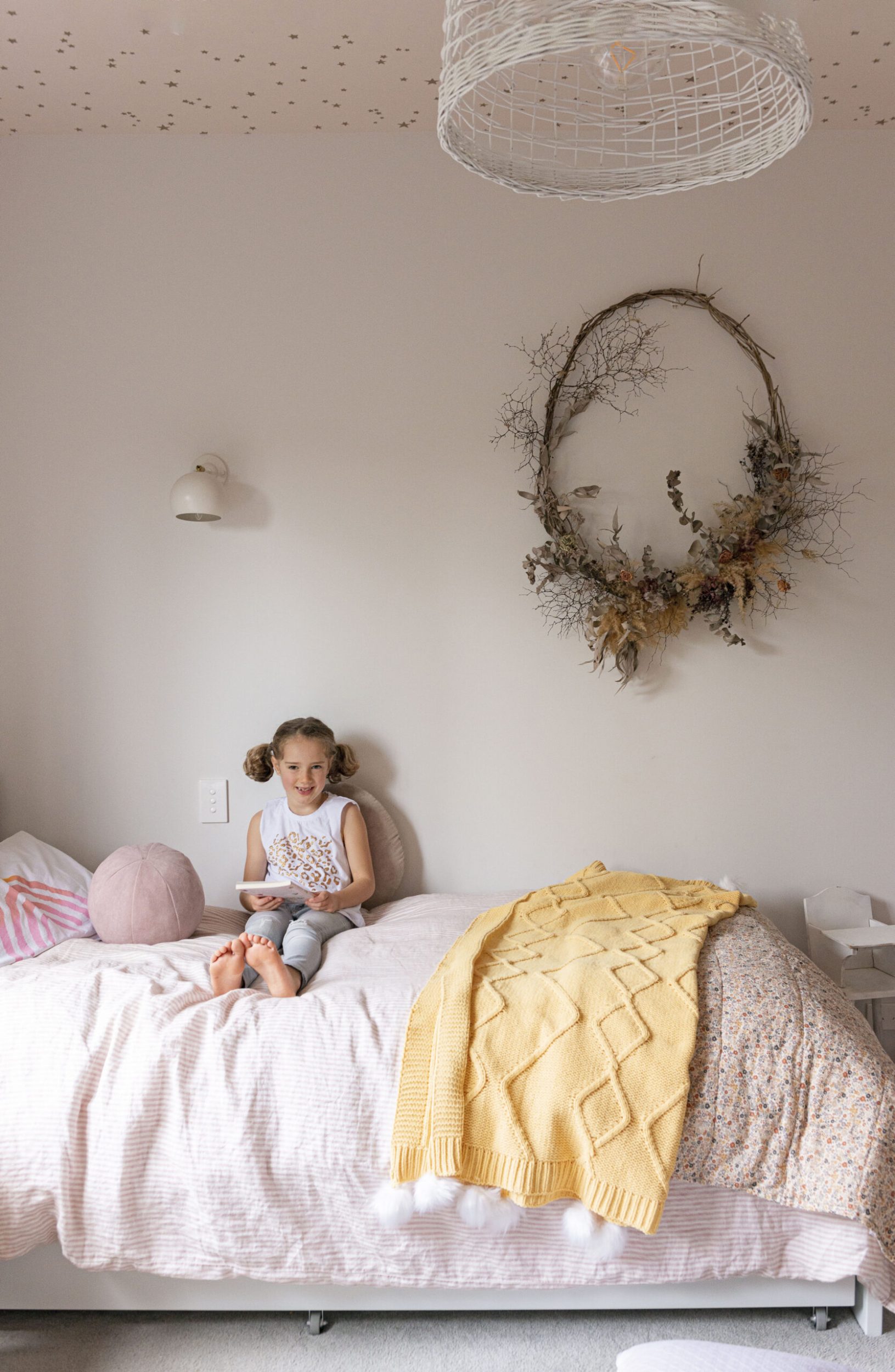 A young girl sitting on her bed with a floral arrangement on the wall and a pastel pink duvet