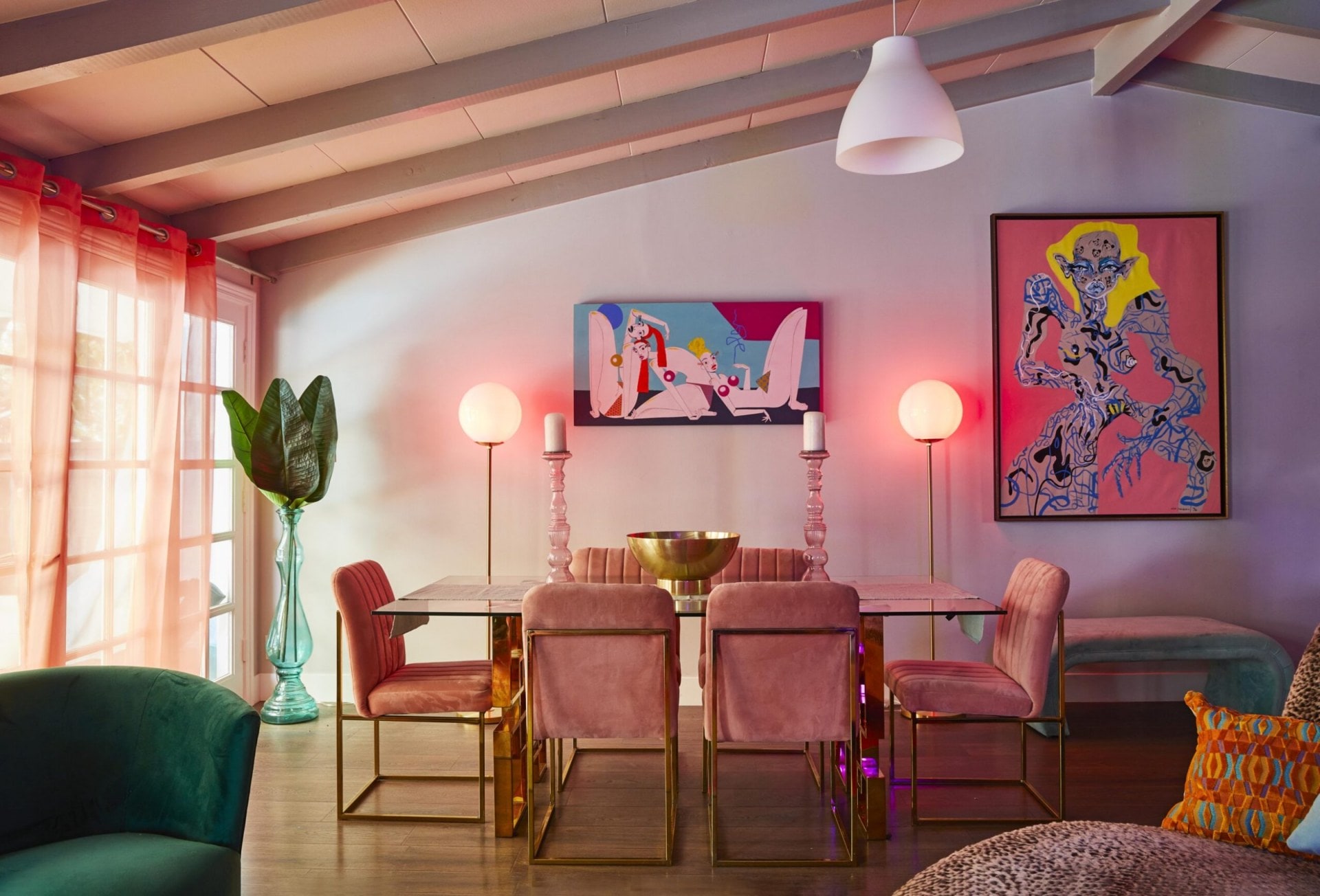Mid century glass dining table with pink chairs, two pink glass candle sticks are on the table and two globe lights are on the back wall with two large pieces of art
