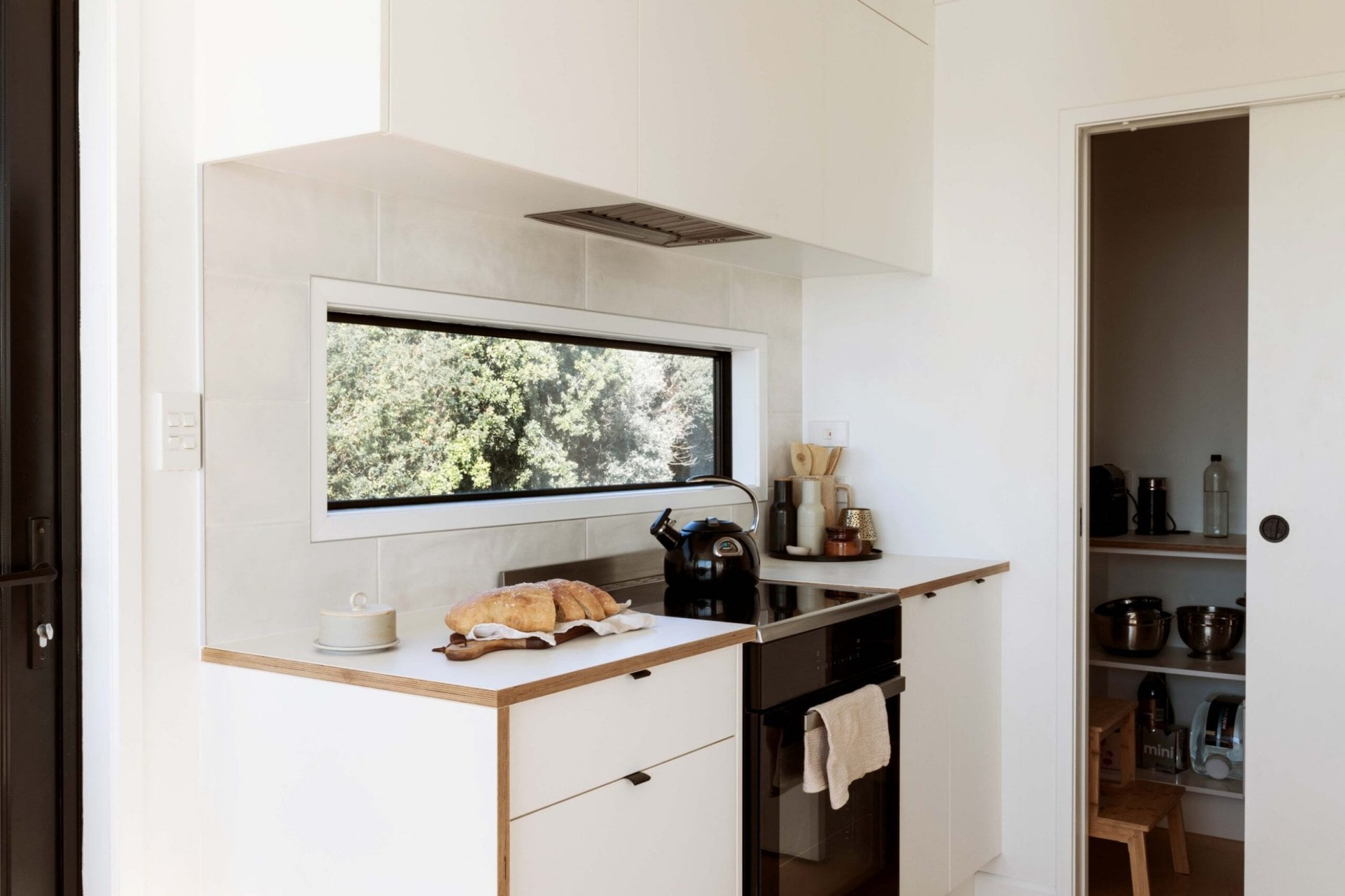 White kitchen cabinets with an oven and window above, with a view to a walk-in pantry