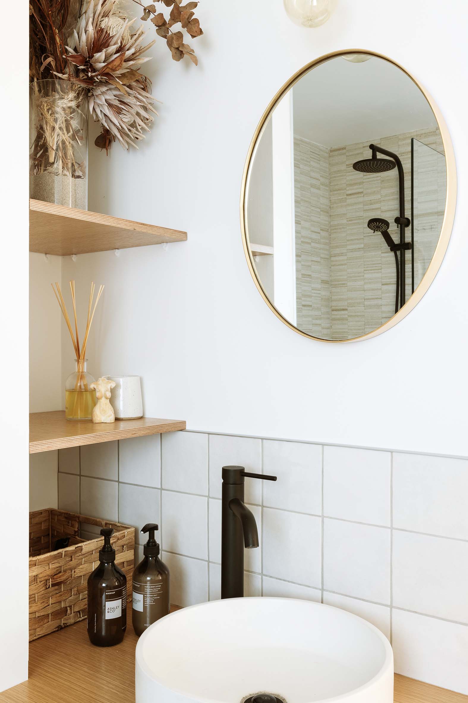 natural bathroom with floating shelves, in the mirror above the white sink and black tap ware you can see the reflection of the shower