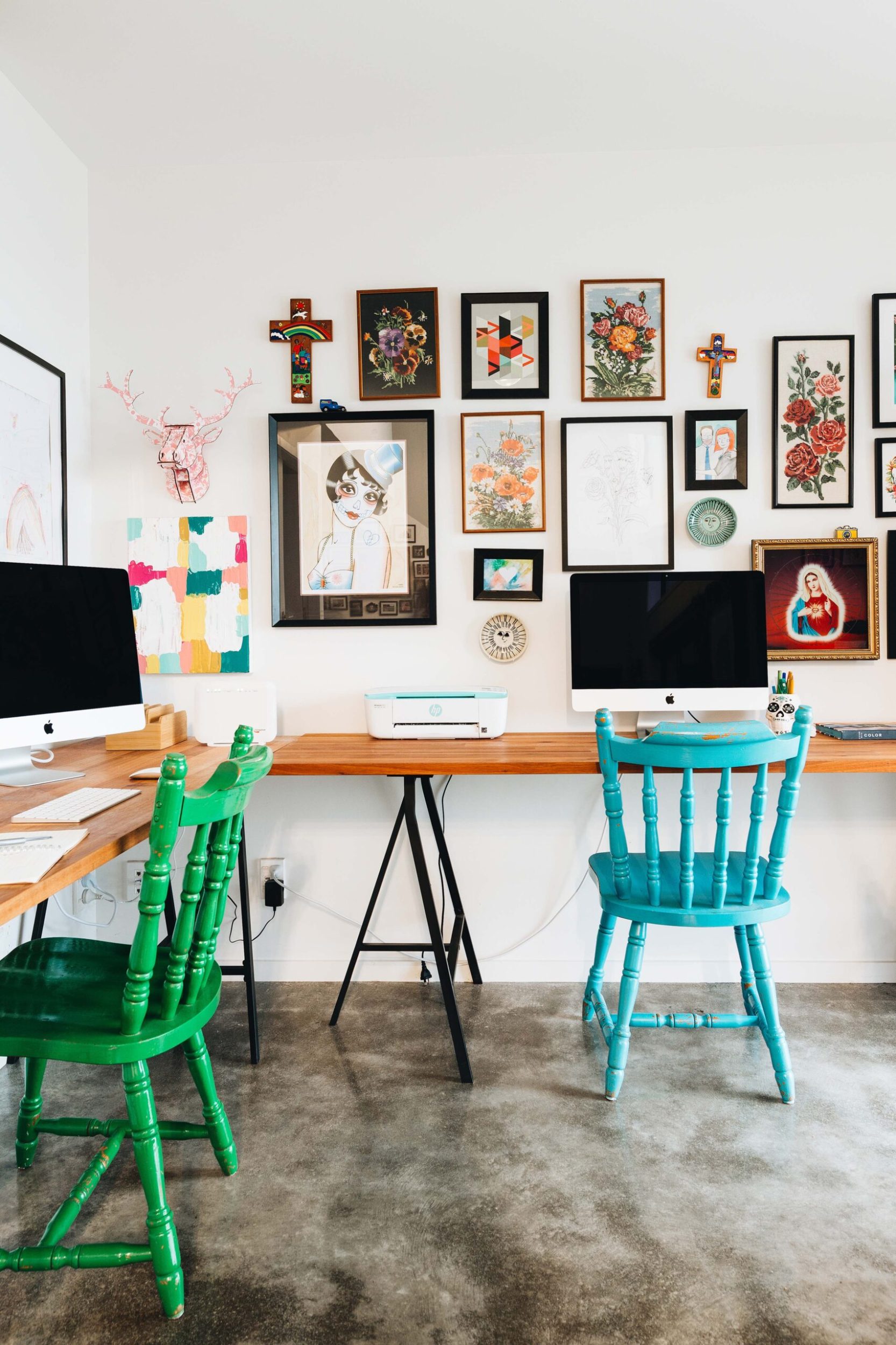 Two desks in a corner with a blue chair and a green chair with various artwork on the wall above the desk