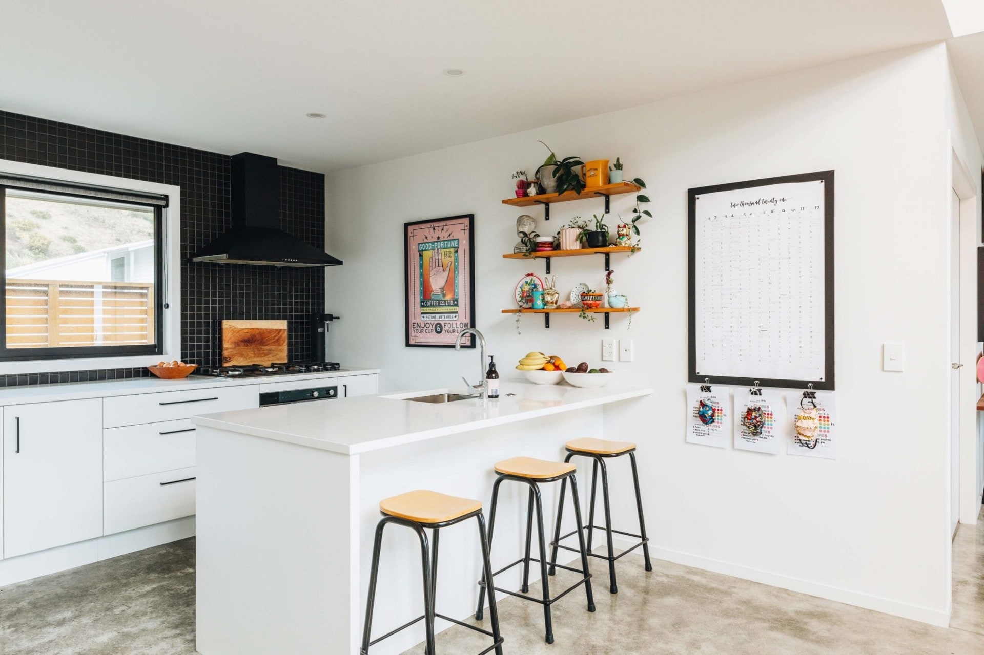 A white kitchen with a black tiled splashback, wooden floating shelves on the wall and a piece of pink artwork, a large calendar hangs on the wall just on the outside of the bench and there are wooden stools at the bench