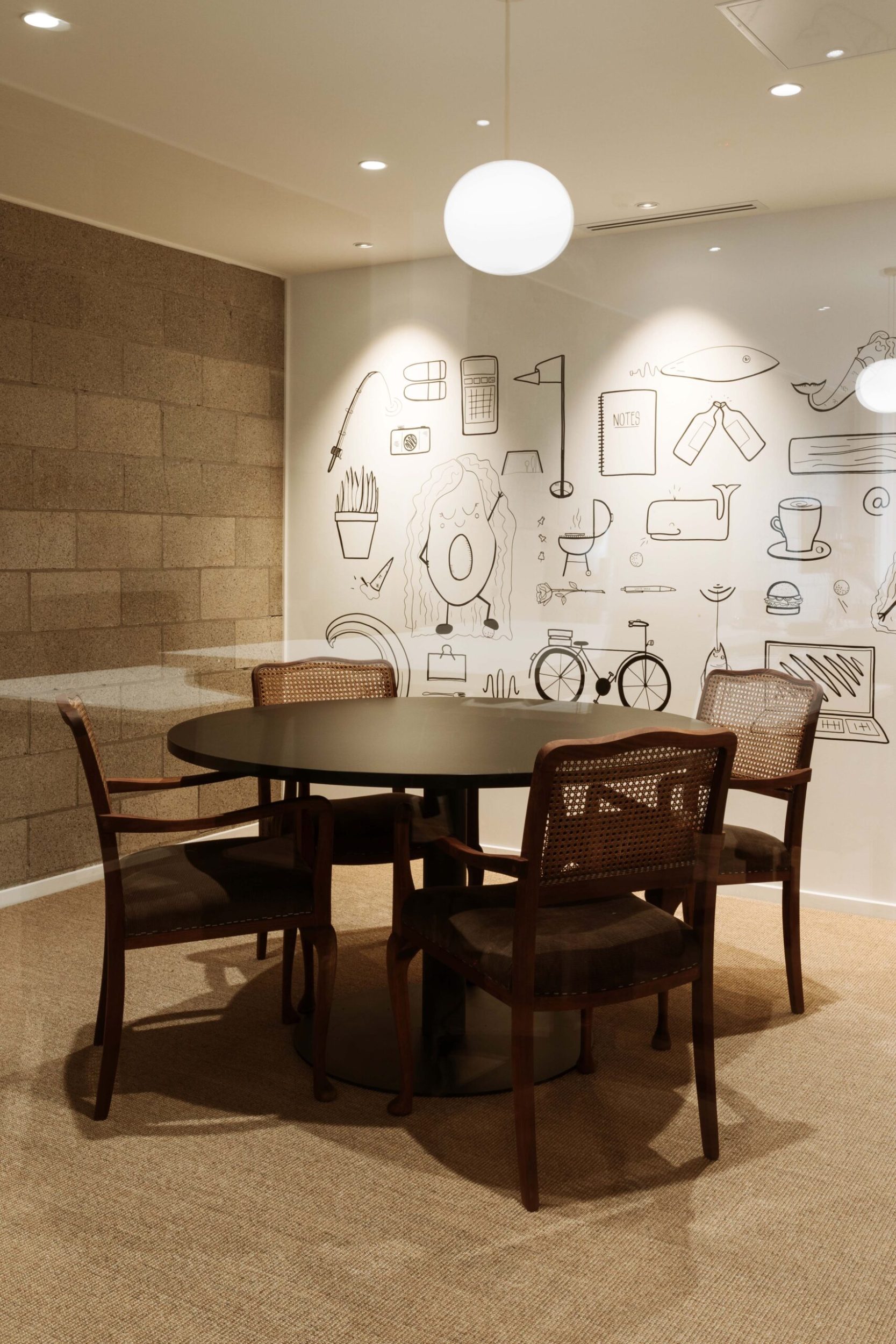 A workspace meeting room with a round table, wooden chairs, a cinderblock wall to the left, and a mural on the back wall