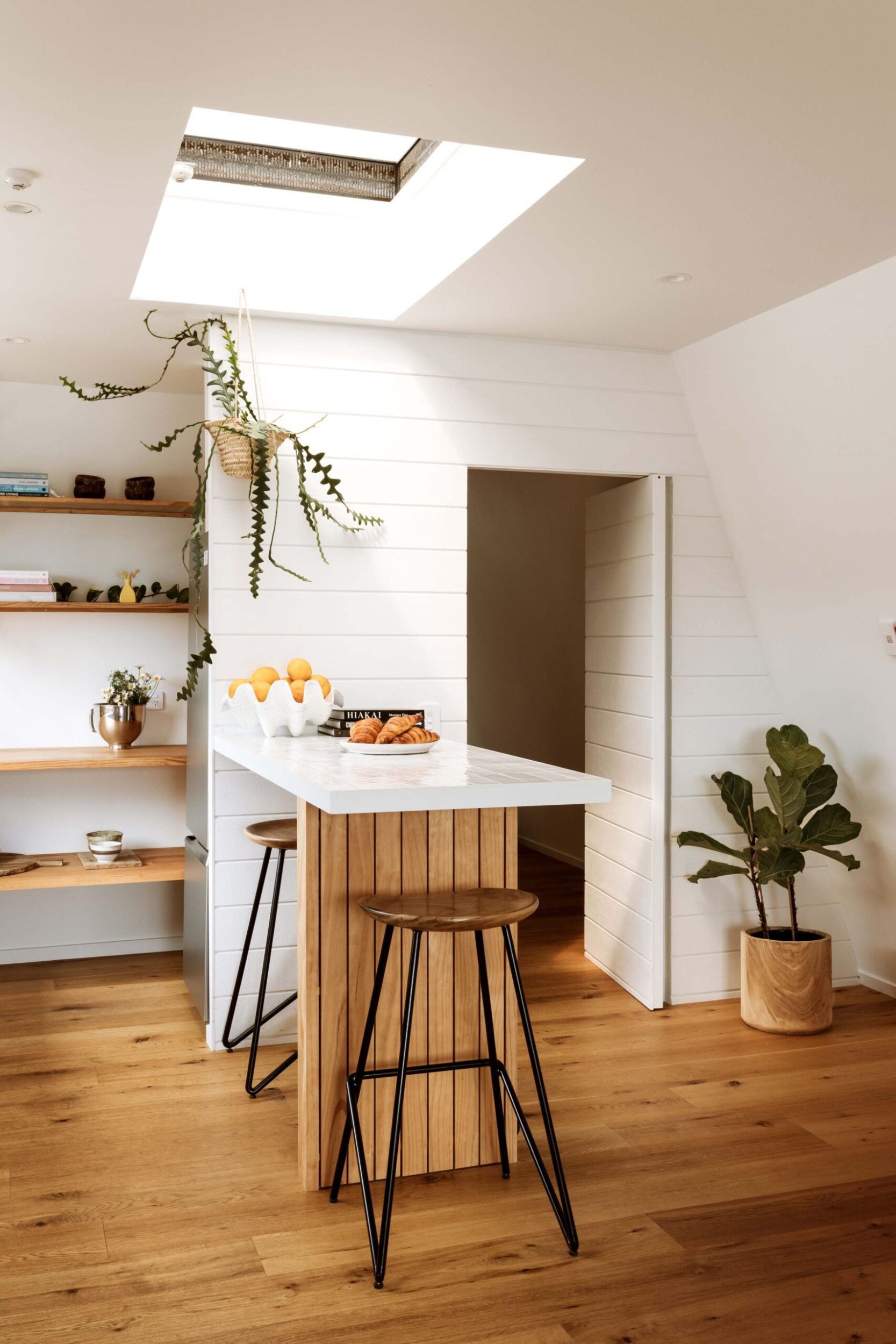 A bright, white kitchen with wooden flooring and panelling up the island, floating wooden shelves are in the corner