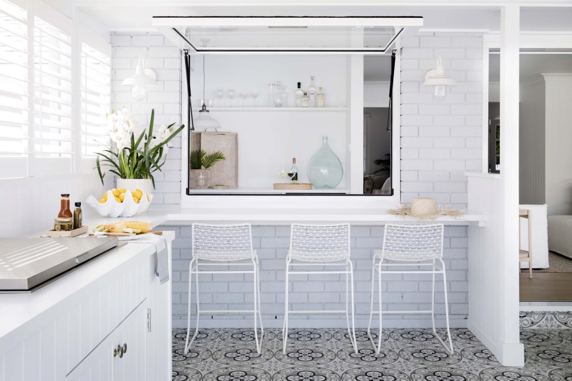 A white outdoor kitchen space with a white benchop and bar stools, a window opens into the kitchen inside