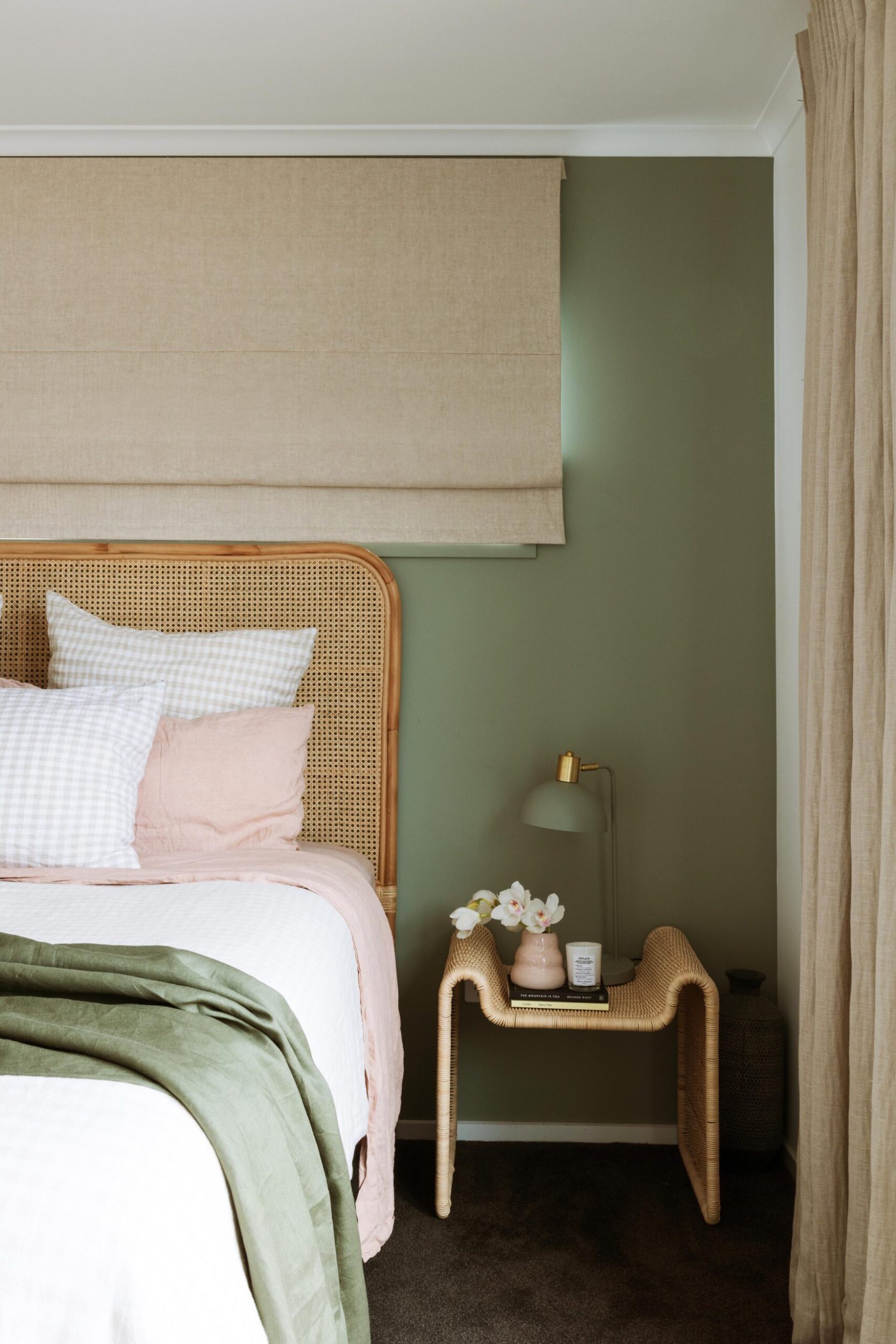 A rattan headboard leaning against a light green wall, a rattan side table and white and blush pink linen dress the bed