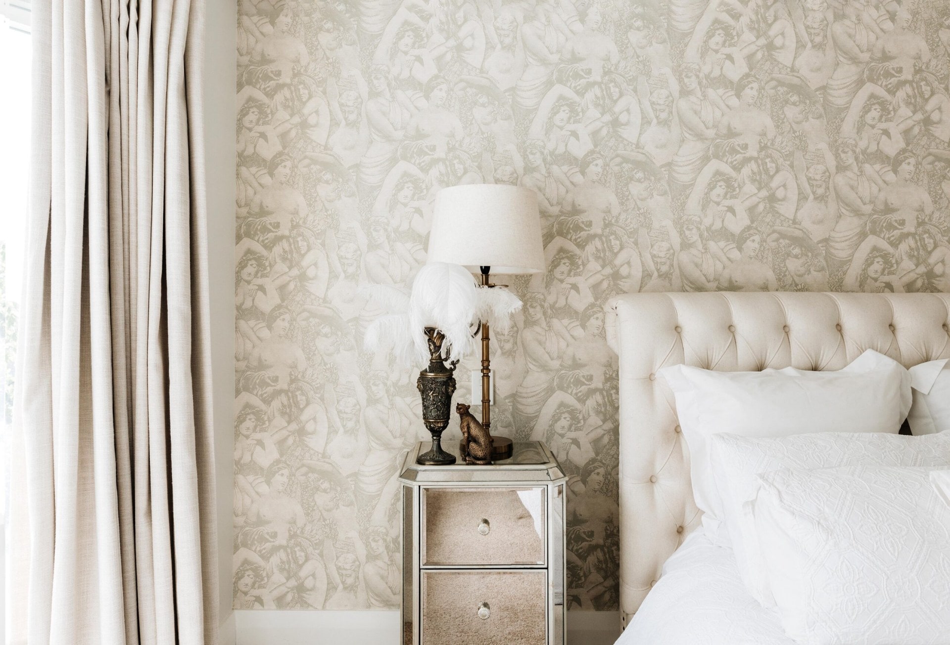 Ornate wallpaper behind a white headboard ,and a glass bedside table