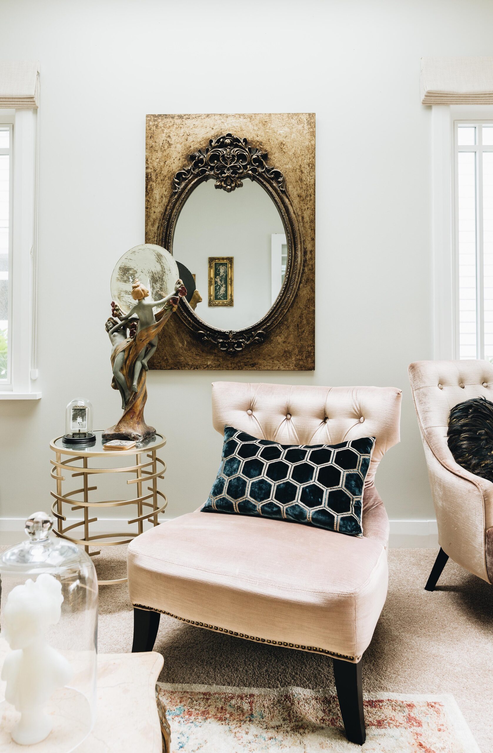 A champagne coloured chair with a black and white cushion next to it is a brass side table with a nymph statue on top