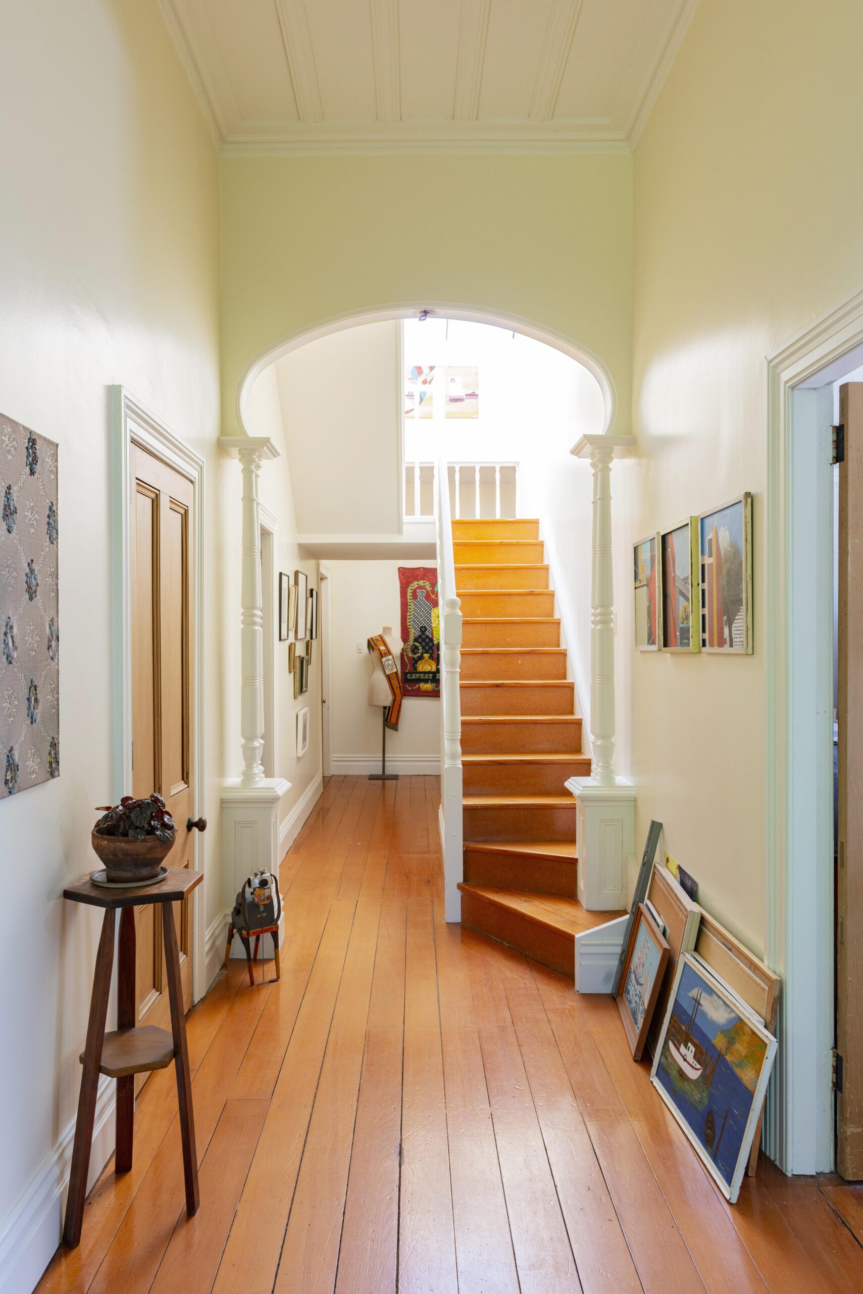 An old villa hallway with floorboard, and a stairwell, there is artwork leaning against the walls and an arch way.