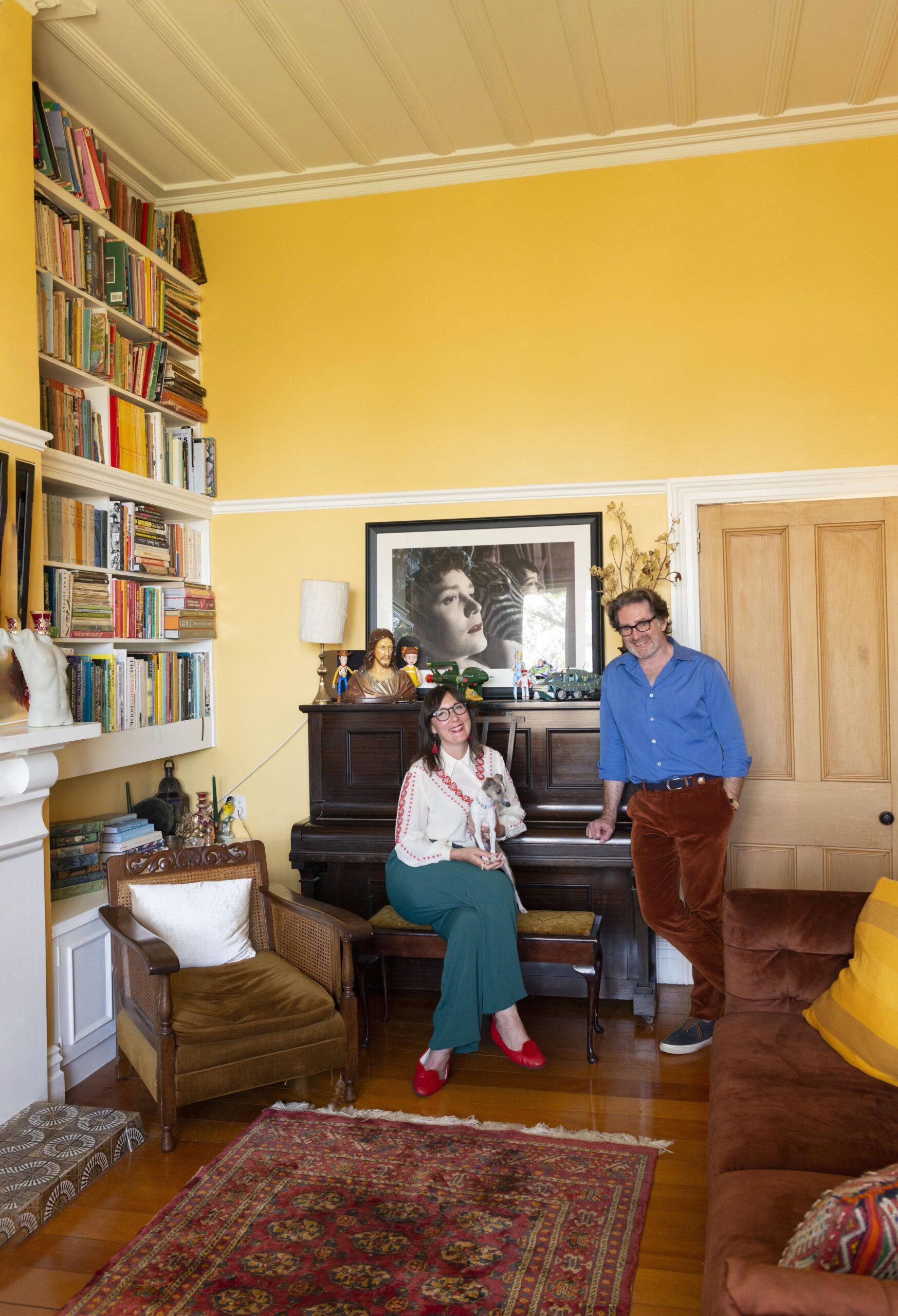 Rose Jackon and Matt Wiseman next to an old piano, the wall behind is a yellow shade, and there is inbuilt shelves stacked with books along the wall