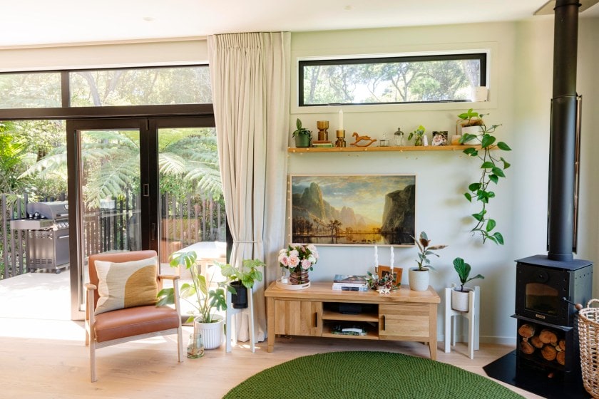 A living room with oak flooring, large windows, a wood burner and tv cabinet, with tv on the wall and a shelf above with a plant hanging down