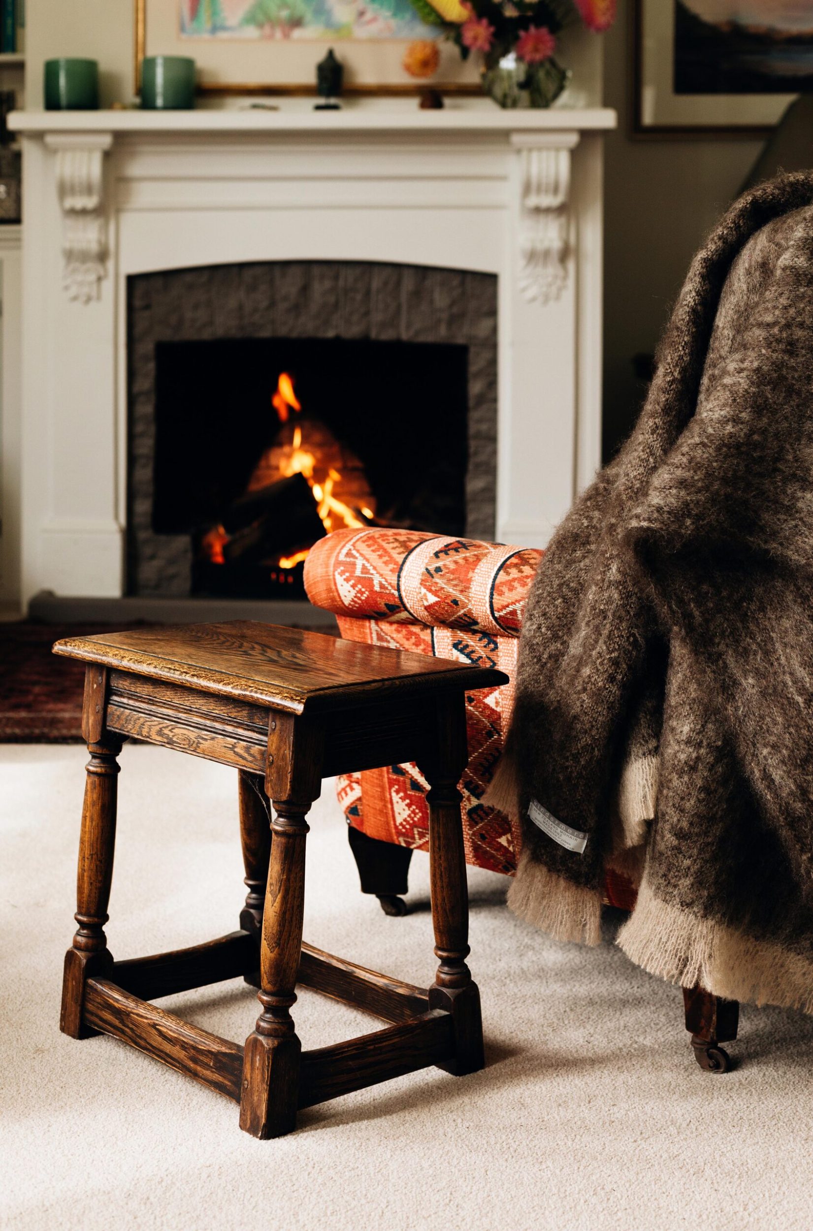 A living room with a white vintage fireplace with the fire lit and a vintage chair and side table in front