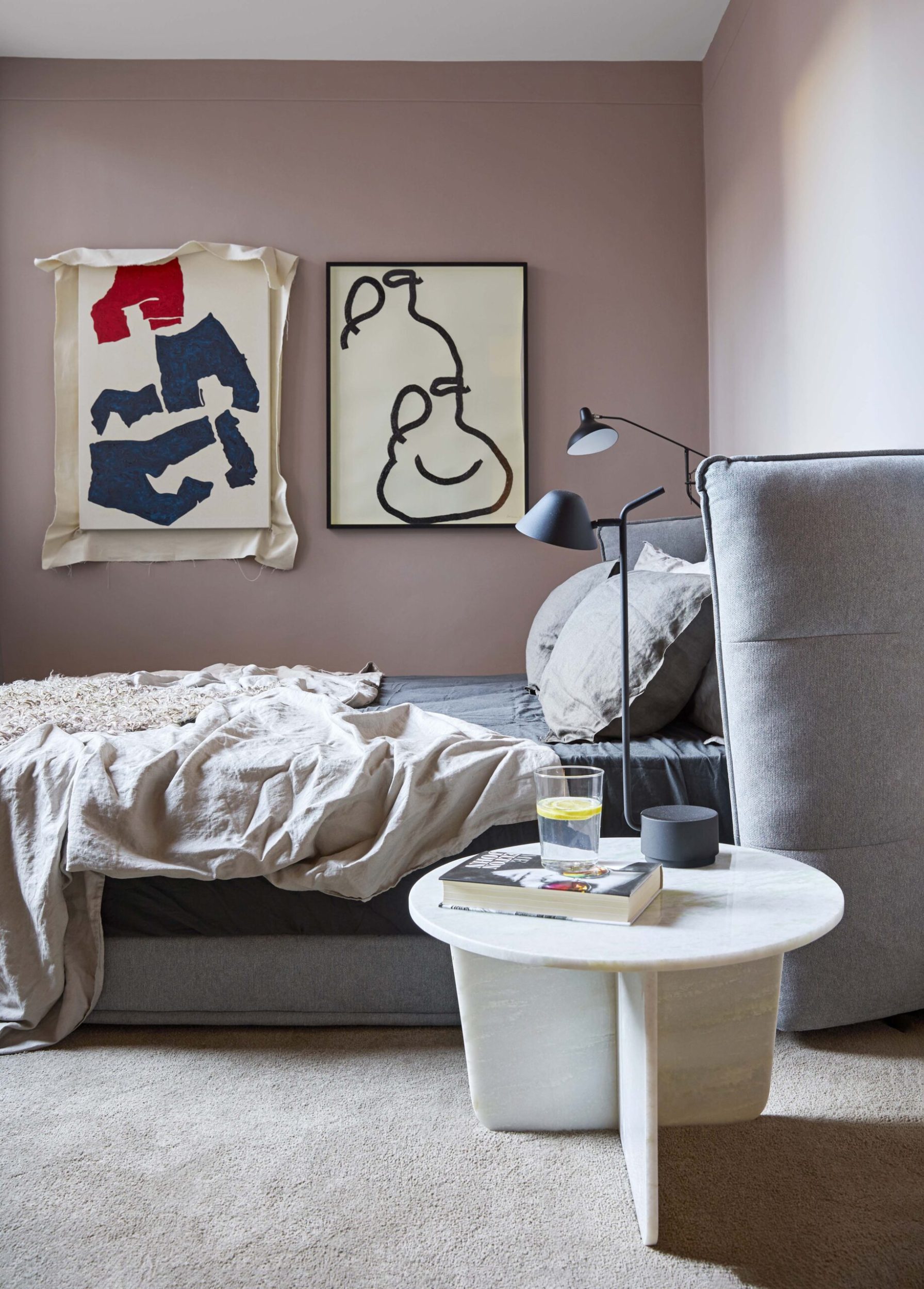 Bedroom with pink walls and a large grey bed with two hanging pieces of abstract art