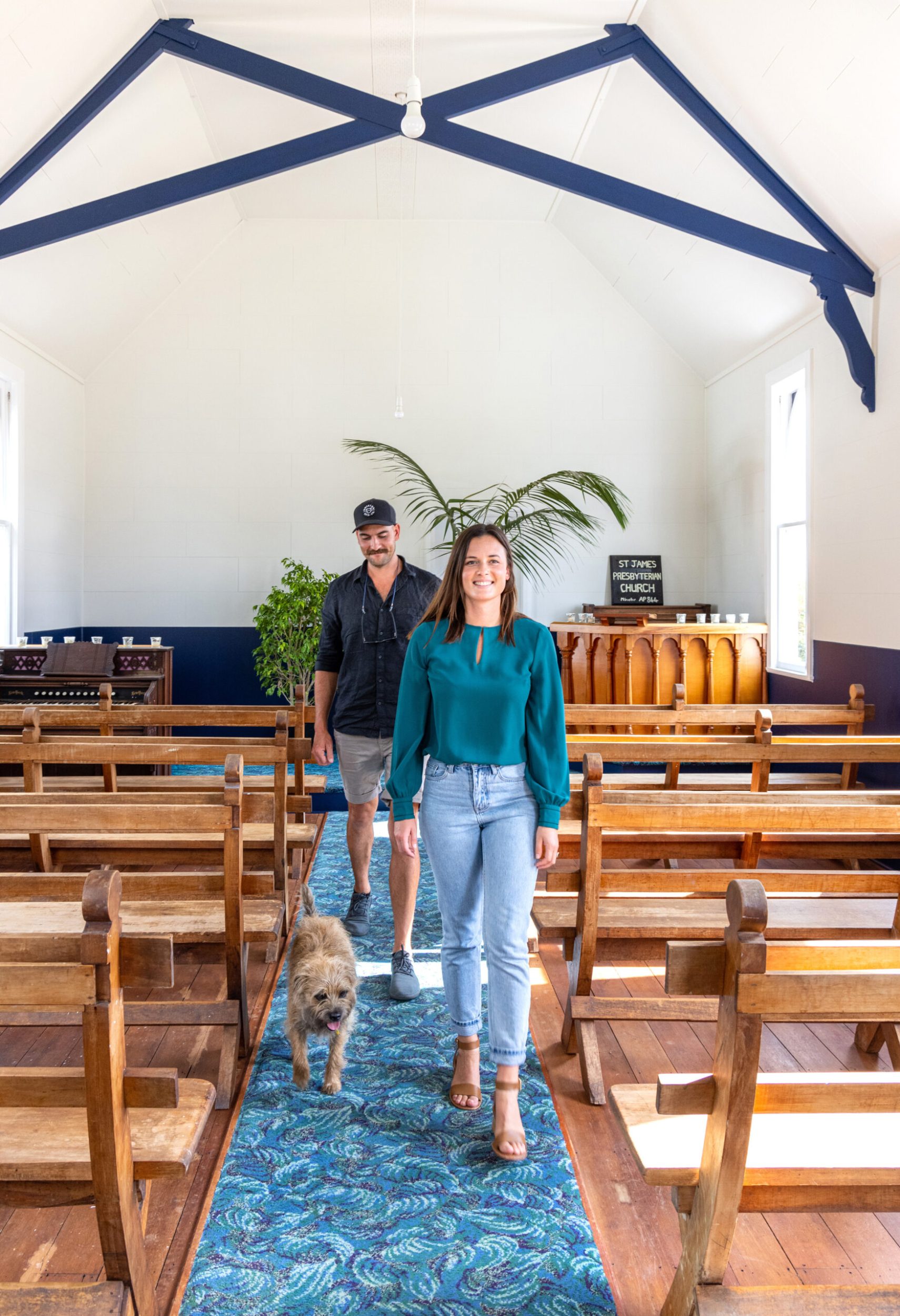 Jemma and Alex Robertson and pet dog Riff Raff walkinginside a white church with brown wooden pews and navy panels