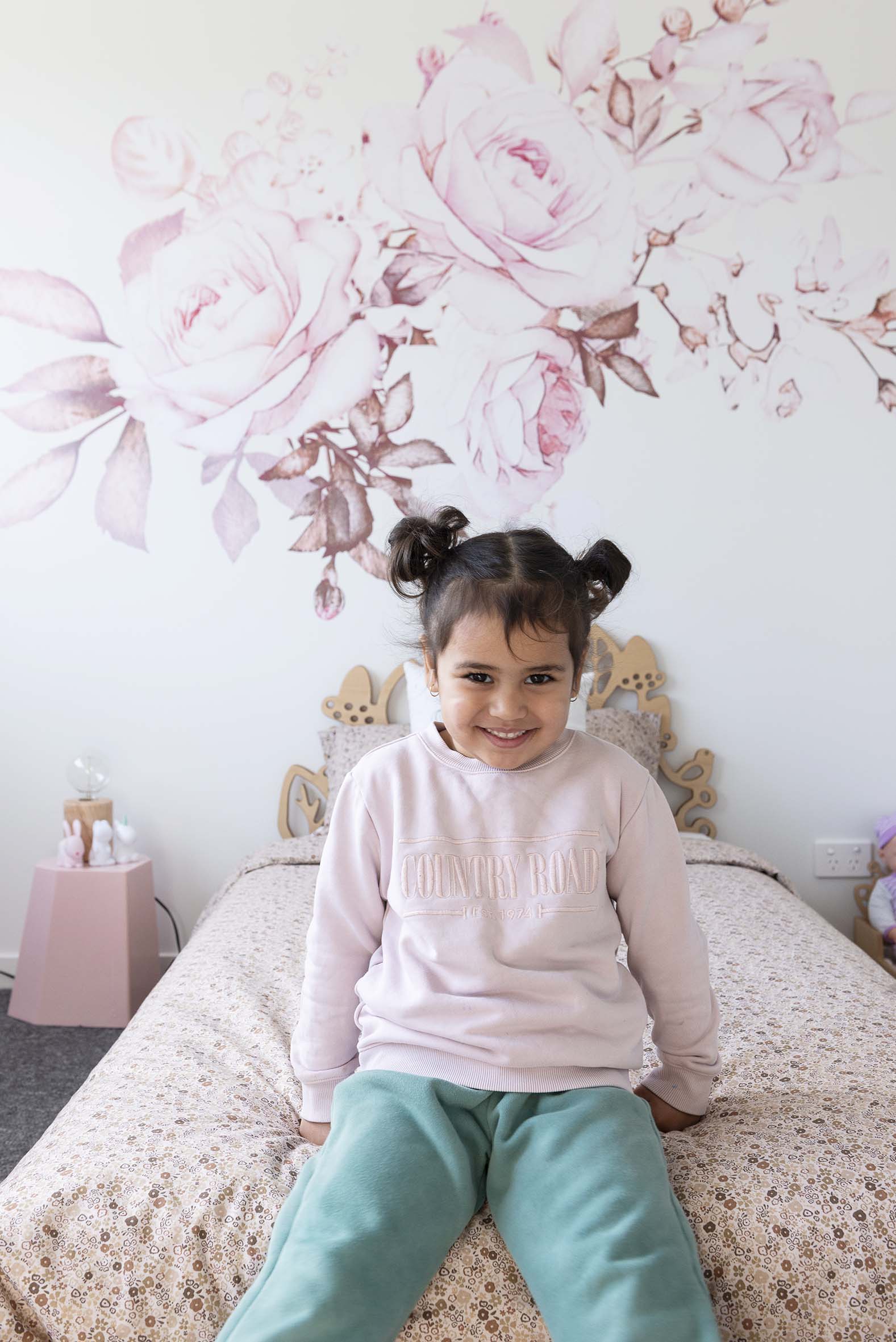 Jude Savea sitting on her bed with pink floral wallpaper on the wall and a pink side table