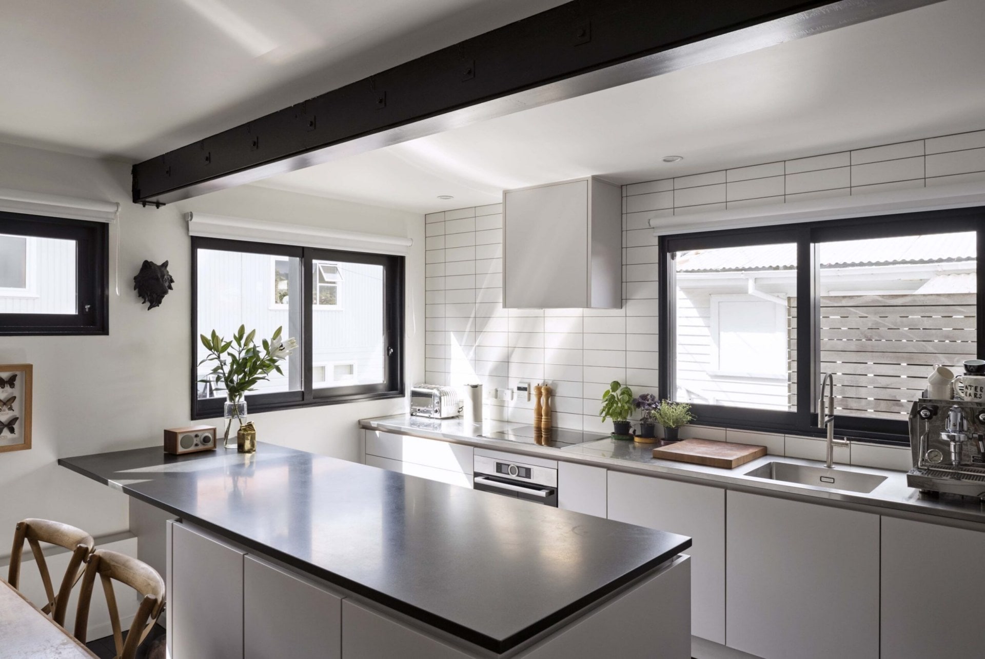 A kitchen with white walls, white drawers with black benchtops, white subway tiles and and a black metal ceiling beam