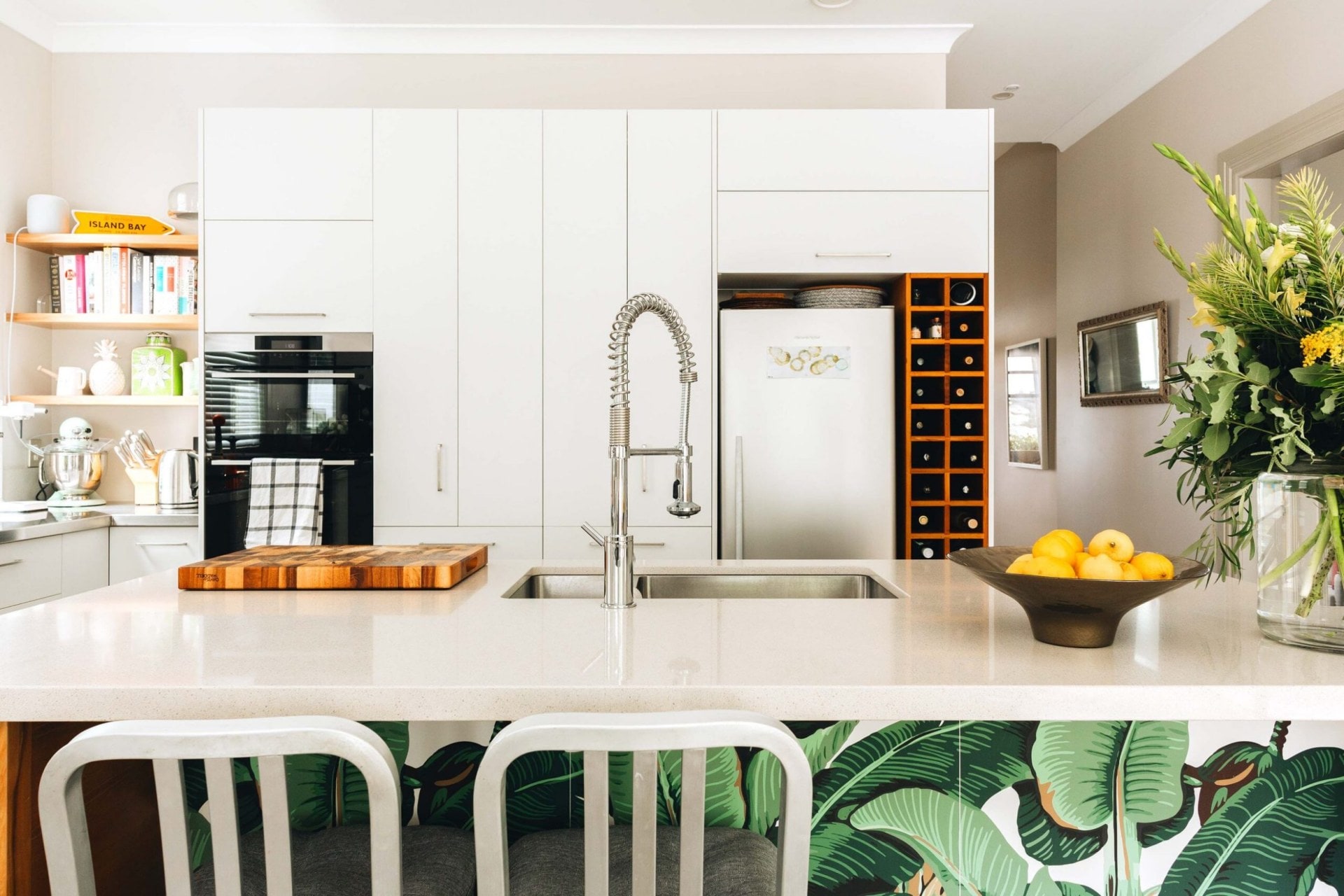 A large white kitchen with white marble benchtops, a silver curved sink tap, a vase of yellow flowers and a botanical splashback