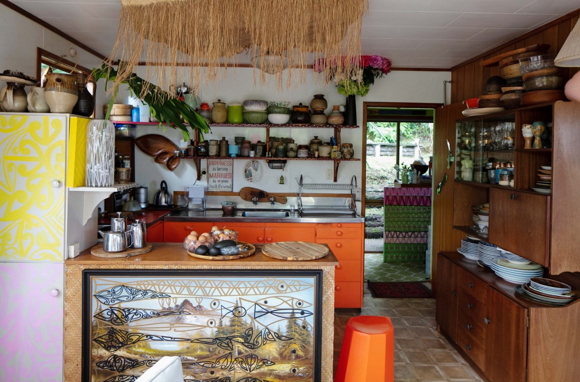 A kitchen with colourful art, orange benches, yellow and purple patterned drawers