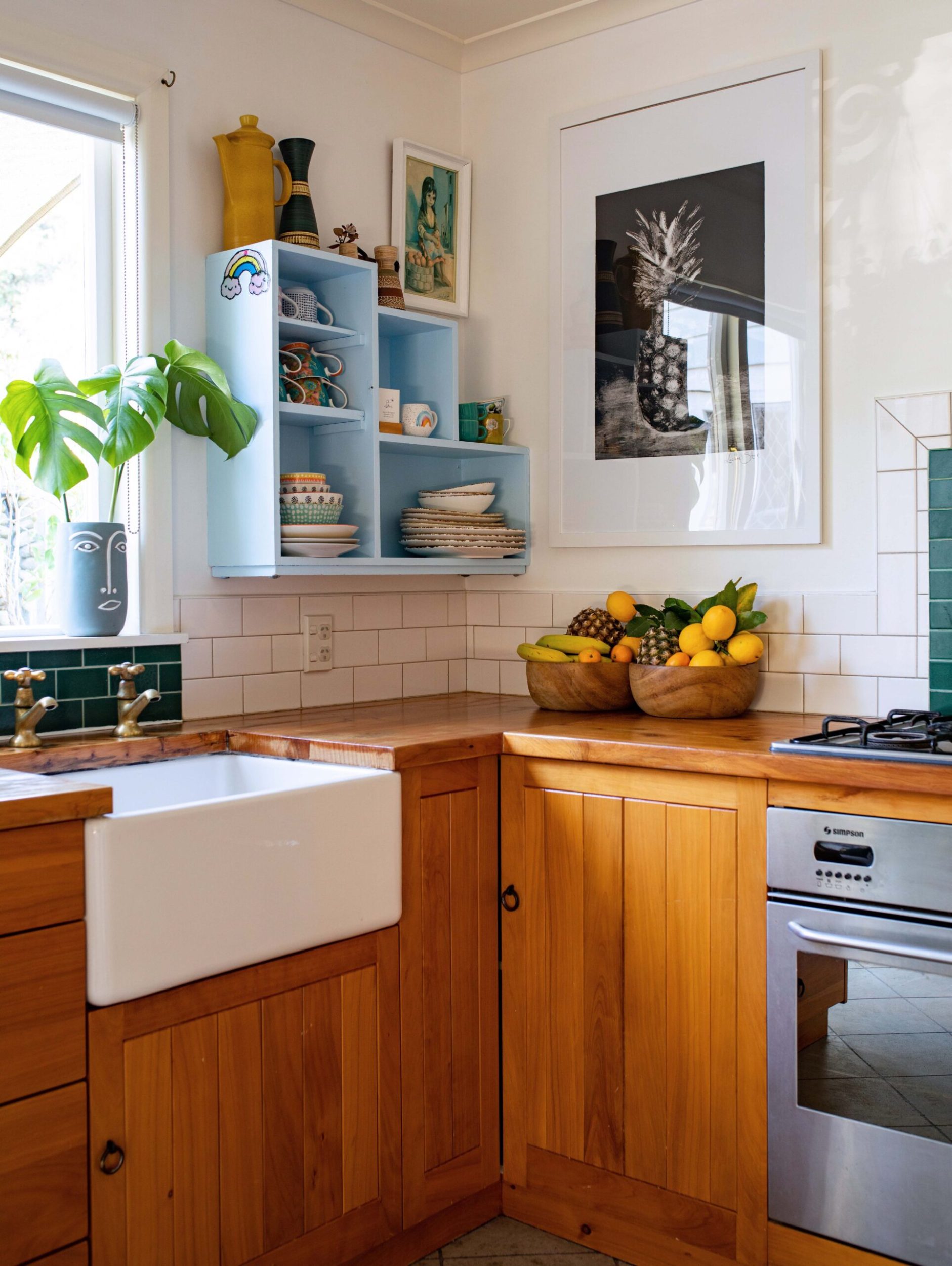 A kitchen with brown wooden cabinets, white floor tiles, a white farmhouse sink and a hanging black and white photo of a pineapple