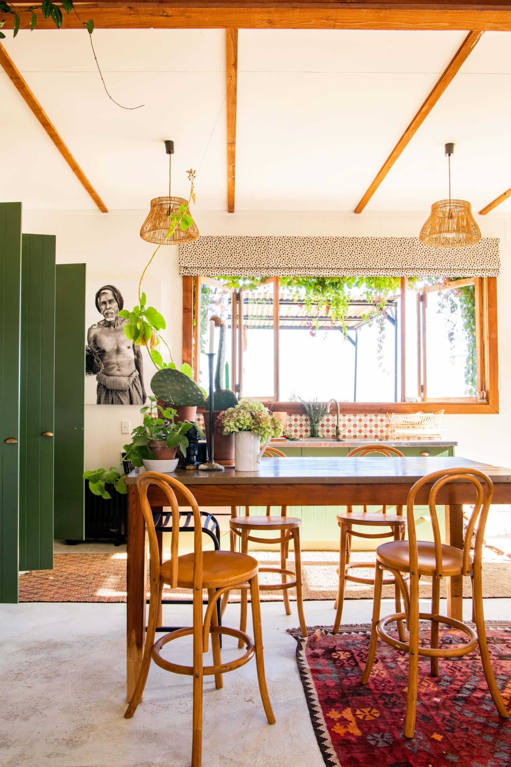 A dining room with wooden beams and window frames, wood tables and chairs and a red boho rug