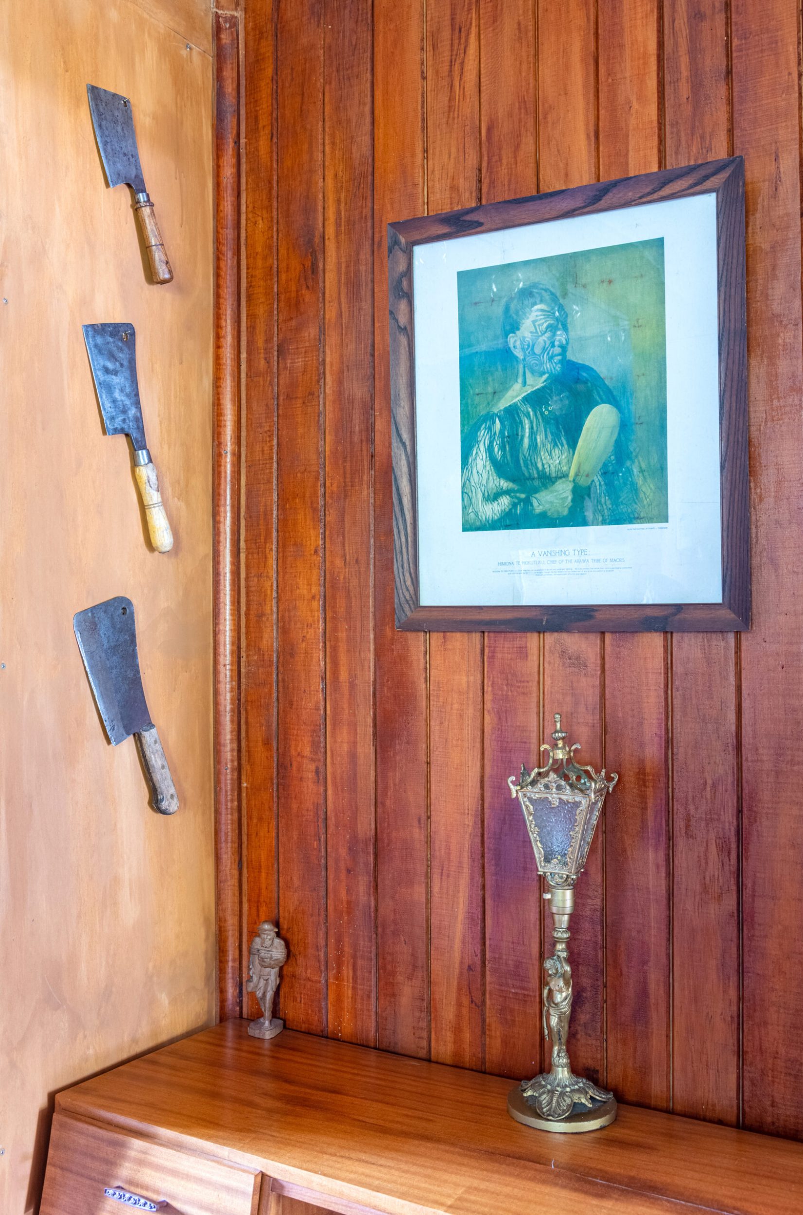 Wood panelled wall with hanging decorated cleaver knives, hanging Māori artwork and a religious ornament