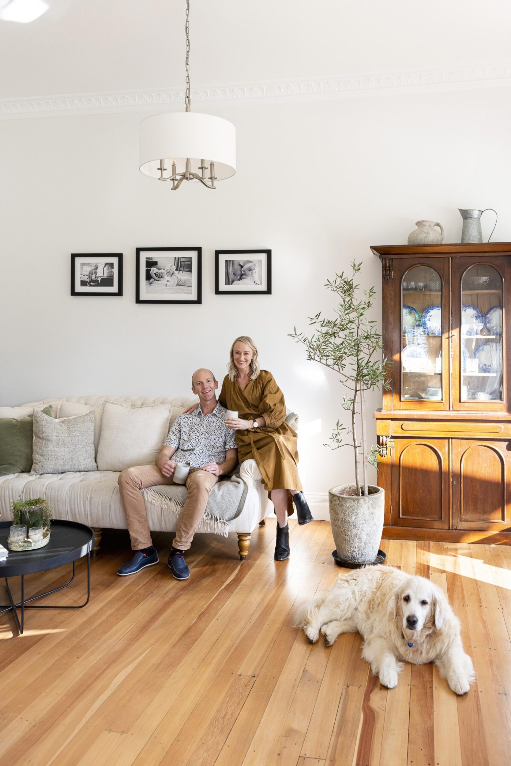 Kylie and John Moody sitting in their living room on a white couch with their white golden retriever Gretel lying on the floor