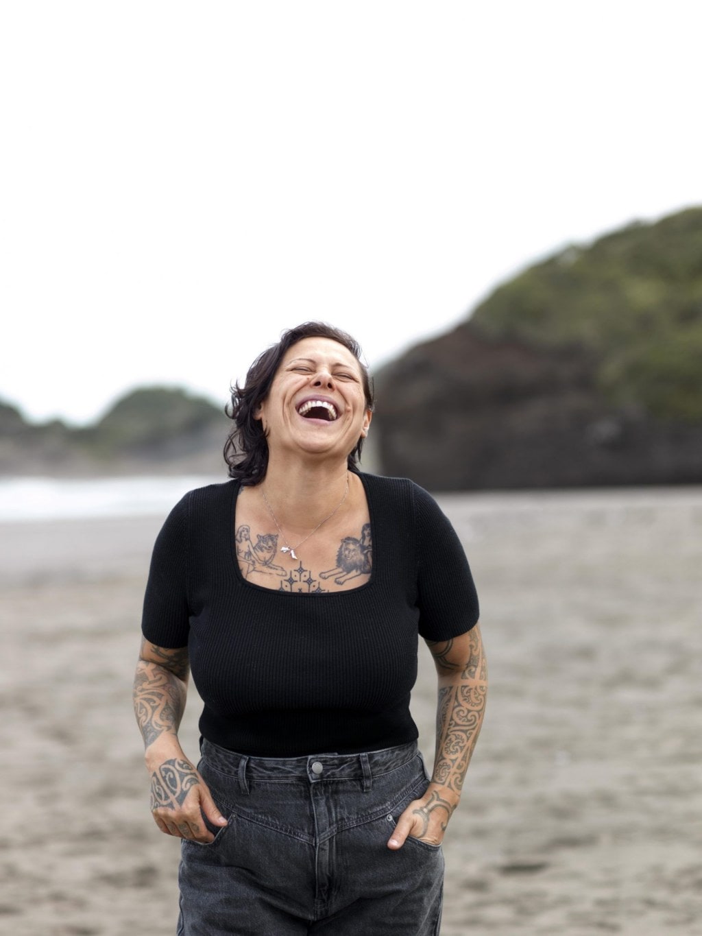 Anika Moa wearing black top and jeans standing at a beach and laughing