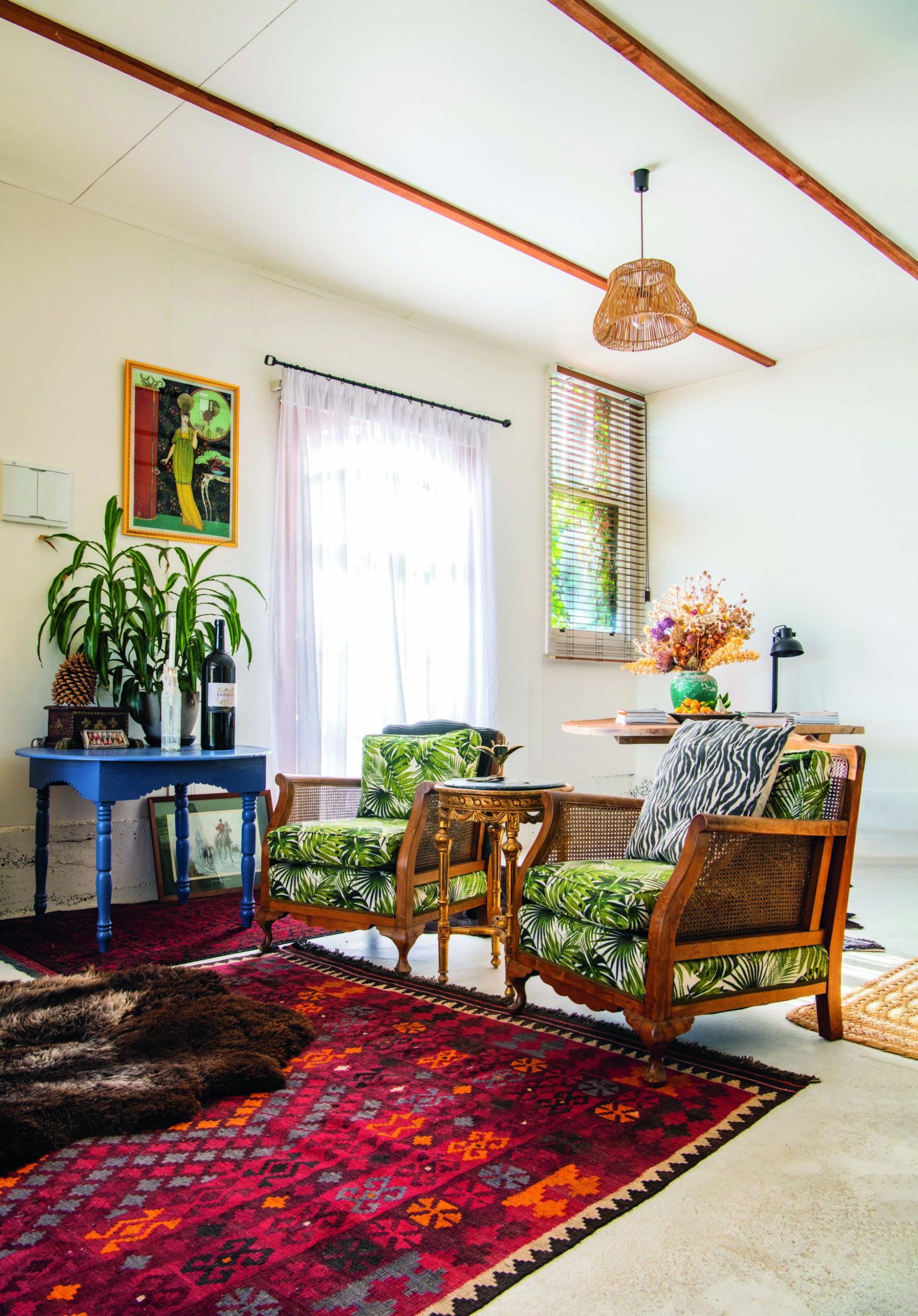 A living room with a red boho rug, green botanical patterned chairs, a blue side table and wooden beams