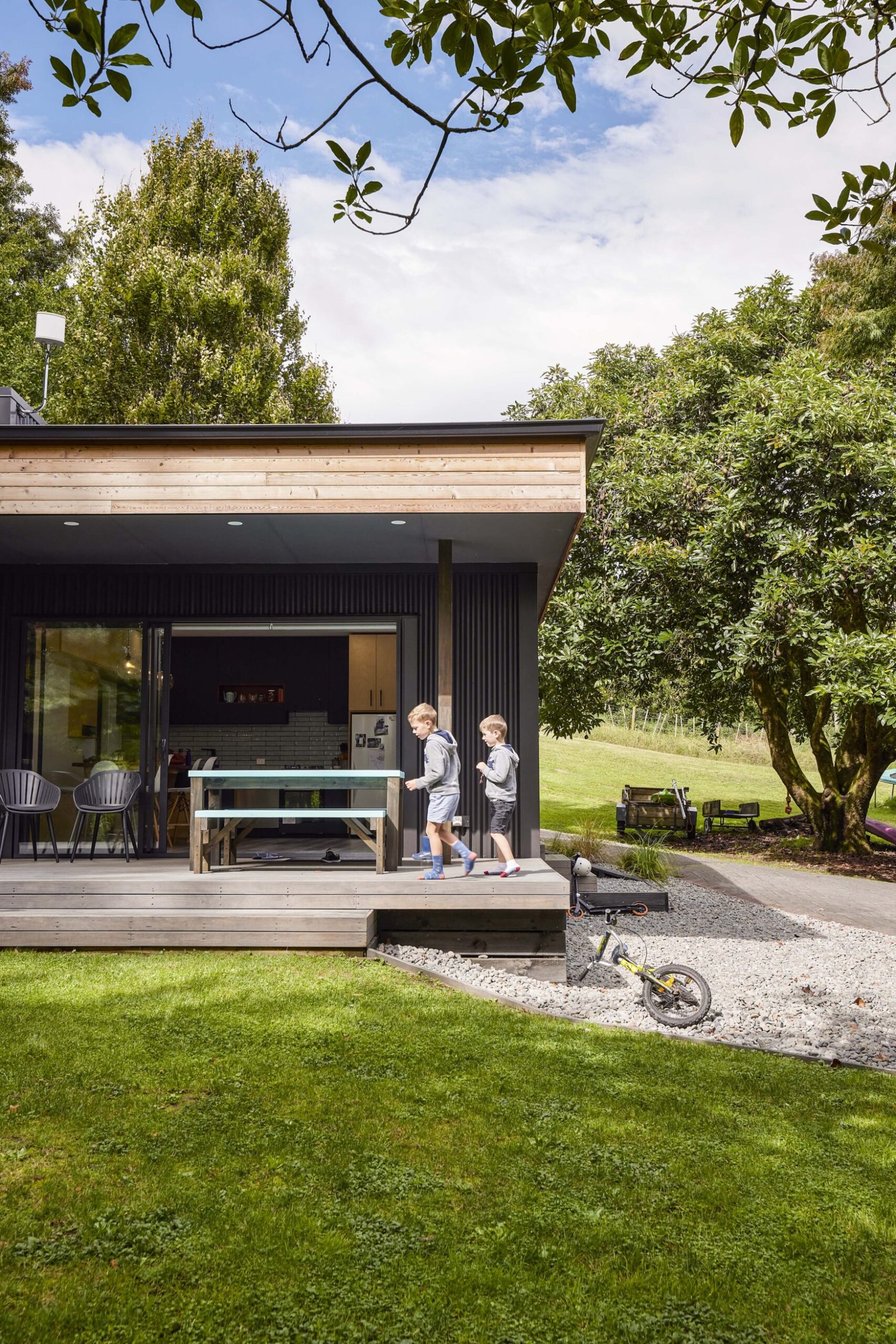 Exterior of a small black home surrounded by nature, with two young boys walking on front porch