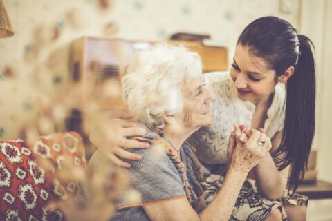 A younger woman with her arm around an elderly woman holding hands