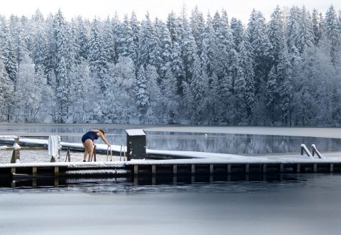 Landscape of a snow-covered forest and lake with a woman wearing a blue swimsuit lowering herself into water